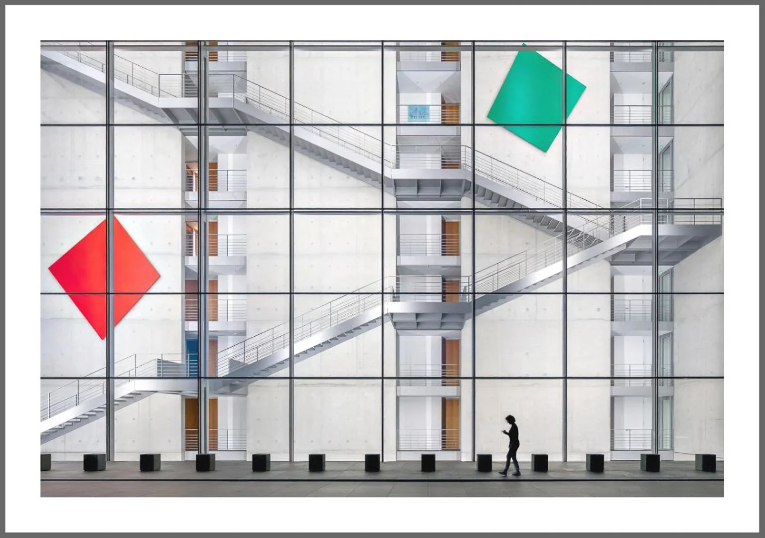 Eine moderne Gebäudefassade mit großen Fenstern, die einen Blick auf ein Treppenhaus im Inneren bieten. Zwei große, bunte Quadrate, rot und grün, sind auf die Fenster geklebt. Im Vordergrund läuft eine Person mit Smartphone, während schwarze Bollard-