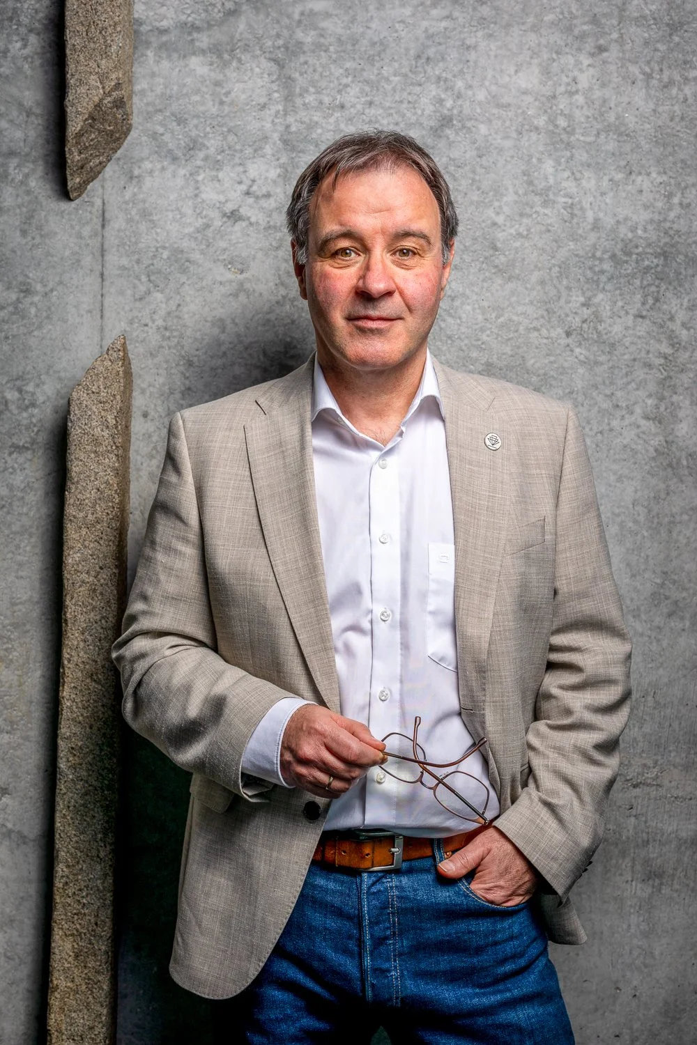 Ein Herr in beigeem Jackett und weißem Hemd steht vor einer grauen Betonwand, hält eine Brille in der Hand und blickt in die Kamera.