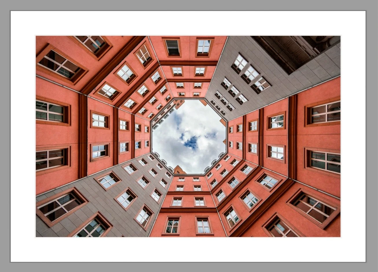 Blick nach oben in einen offenen Innenhof eines mehrstöckigen Gebäudes mit roten und grauen Fassaden, Fenstern und Himmel sichtbar in der Mitte.