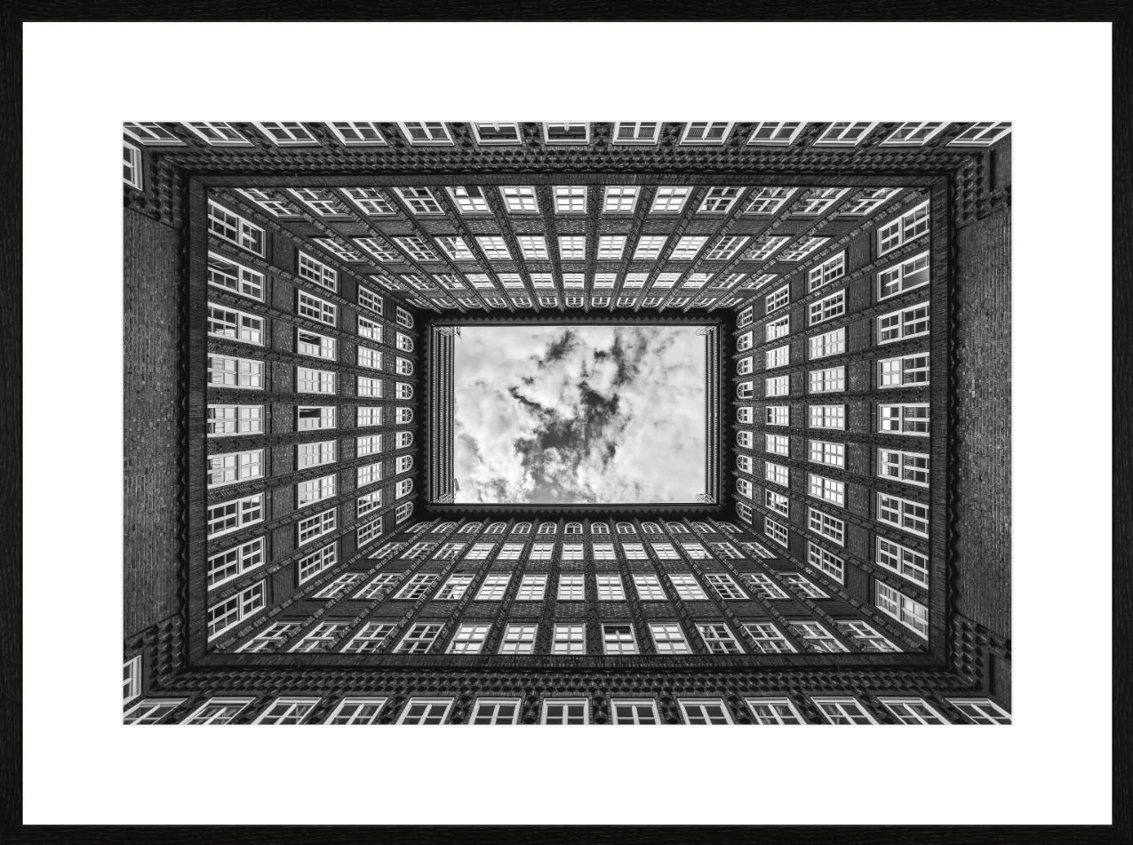 Blick nach oben in einen quadratischen Innenhof eines mehrstöckigen Gebäudes mit Fenstern, Blick auf den Himmel mit Wolken.