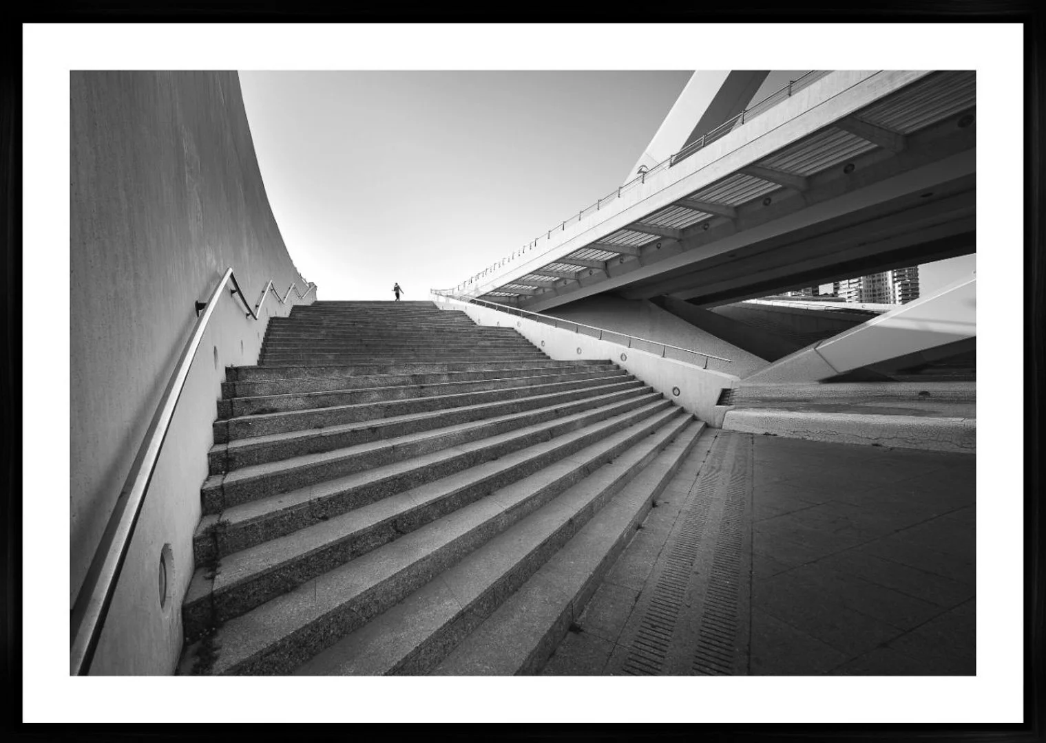 Schwarz-weiße Aufnahme einer modernen architektonischen Struktur mit breiten Treppen, die nach oben führen, und einer Person, die oben auf den Stufen steht. Große Gebäude und Himmel im Hintergrund.