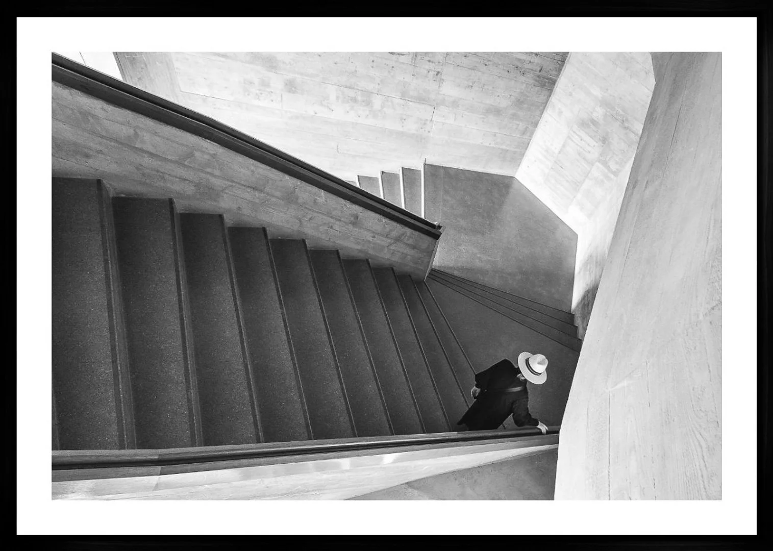 Schwarze Treppe in einem modernen Gebäude, eine Person mit schwarzem Mantel und Strohhut geht die Treppe hinunter.