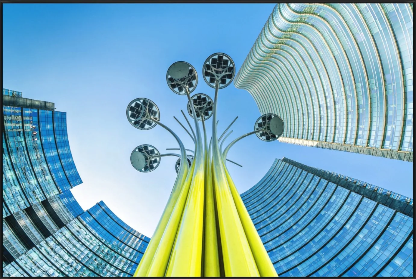 Blick nach oben auf eine moderne Stadt mit Hochhäusern und einer Kunstinstallation aus gelben Säulen und reflektierenden runden Elementen gegen den blauen Himmel.