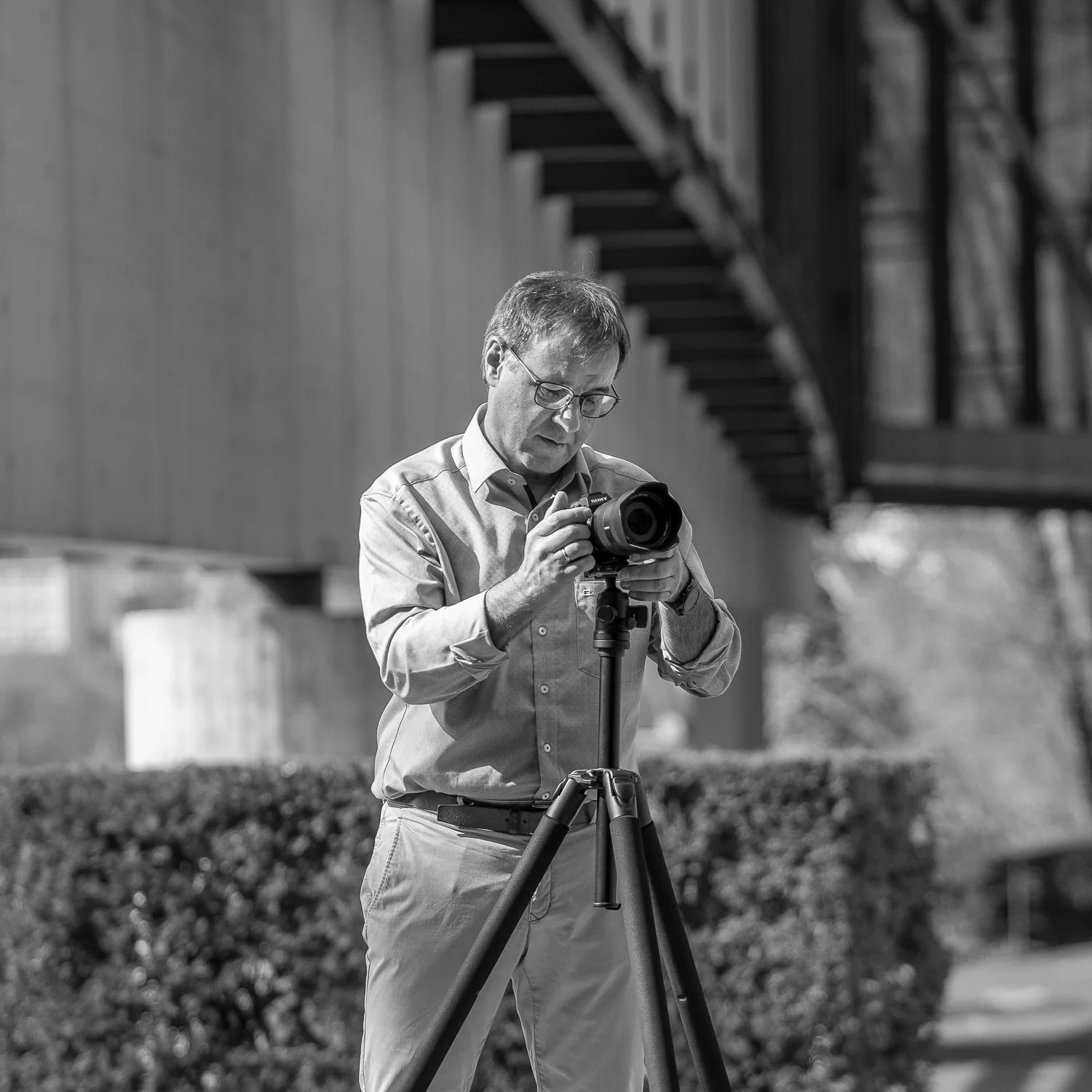 Mann steht vor Busch, benutzt Kamera auf Stativ, unterwegs fotografieren, monochrome Aufnahme
