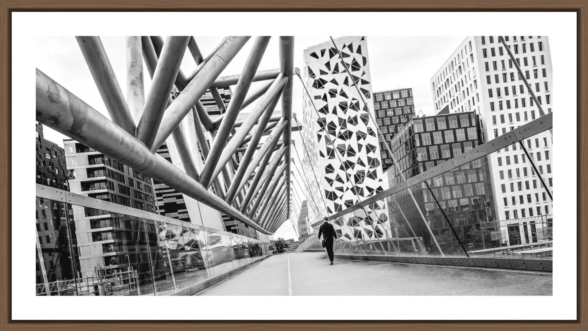 Schwarz-Weiß-Foto einer modernen städtischen Brücke mit architektonisch auffälligem Design, umgeben von hohen Gebäuden, eine Person läuft die Brücke entlang.