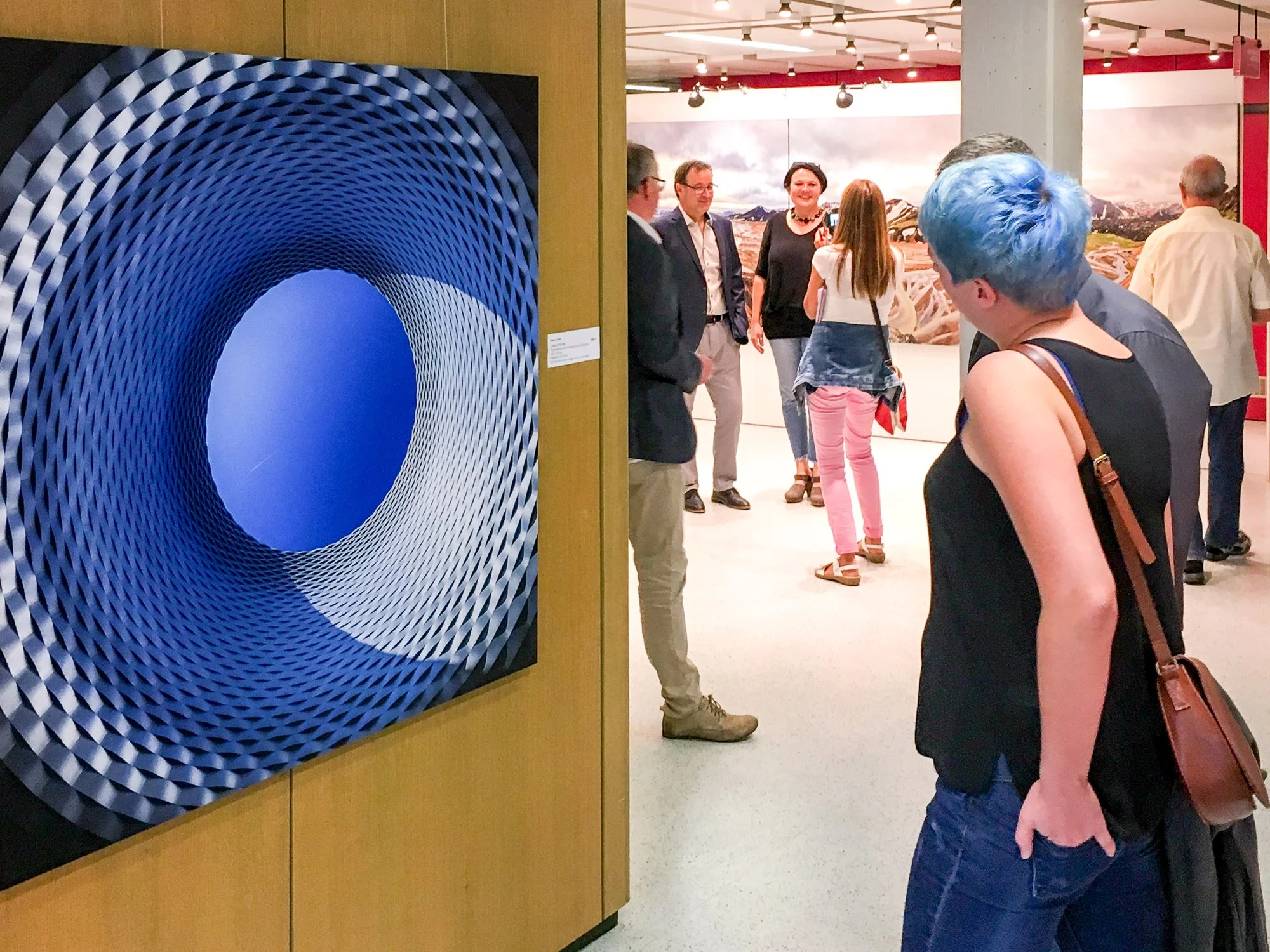 Kunstwerk mit einem blauen, geometrischen, hohlzylindrischen Design, das in einem Kunstmuseum ausgestellt ist, während Besucher das Bild betrachten.