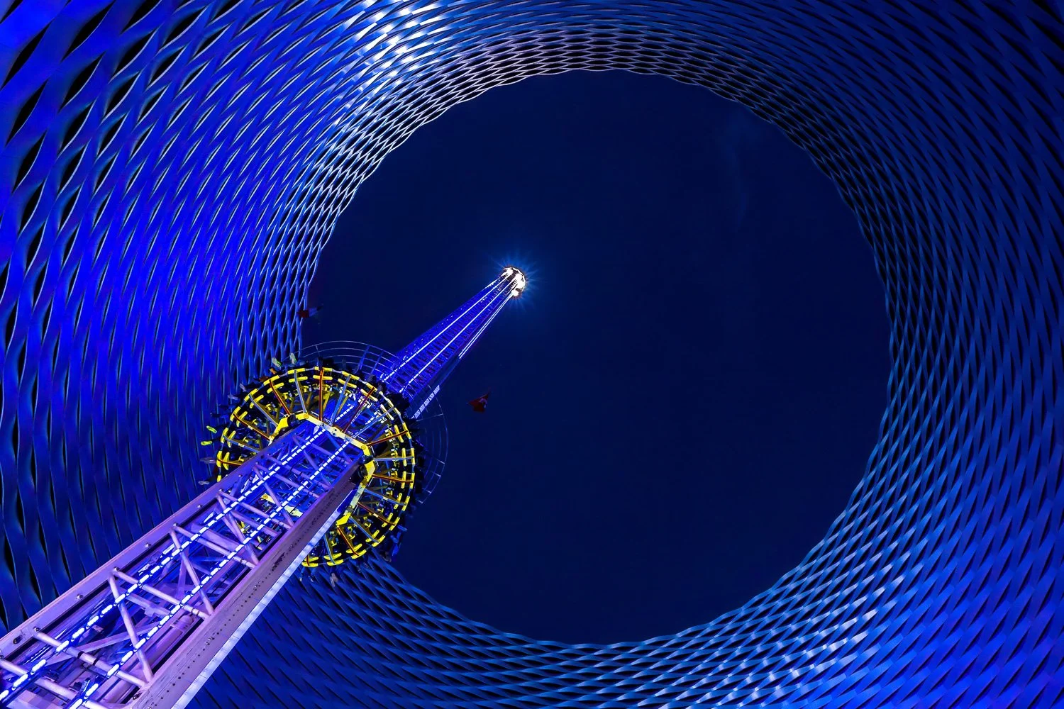Blick nach oben auf ein Riesenrad bei Nacht, umgeben von einer blauen, metallischen, spiralförmigen Struktur.