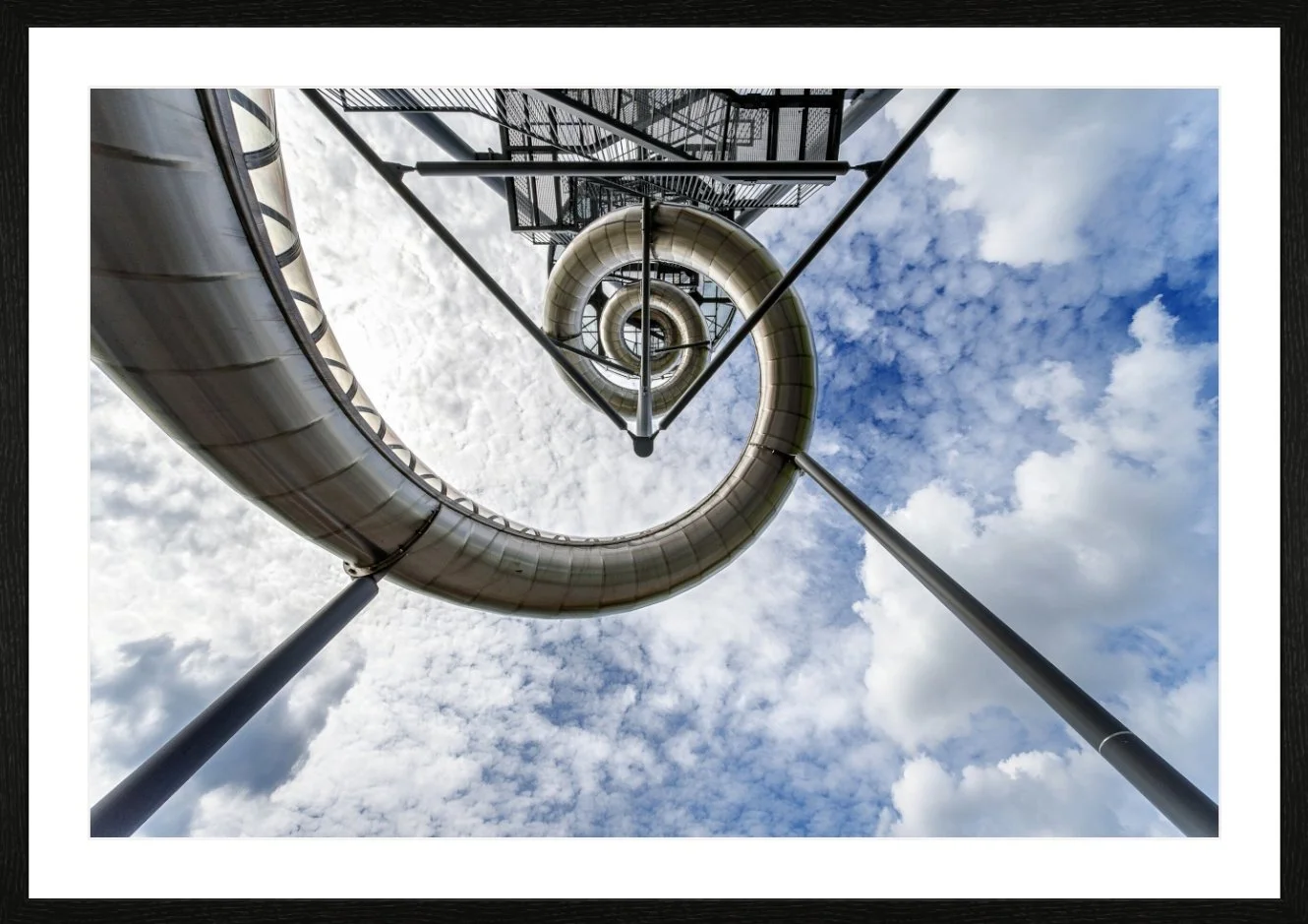 Blick nach oben auf eine spiralförmige Rutsche auf einem Spielplatz oder Freizeitpark unter einem bewölkten Himmel.