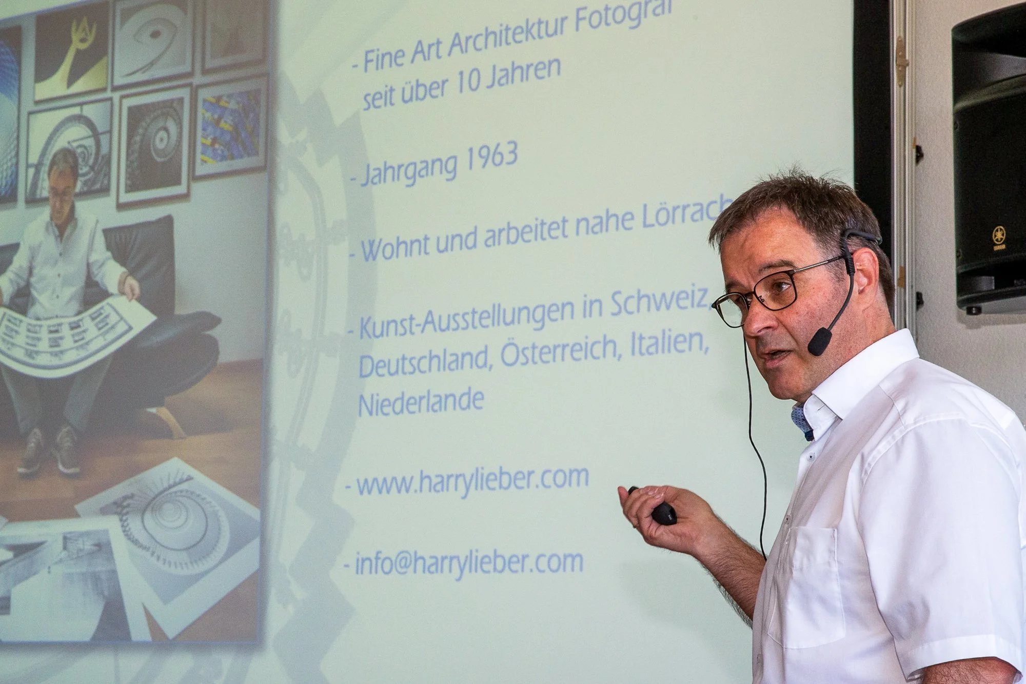 Ein Mann mit Brille und Headset hält einen Vortrag vor einer Präsentation. Auf dem Bildschirm sind Textinformationen über Kunst und Architektur sowie Kontaktdaten und eine Website zu sehen.
