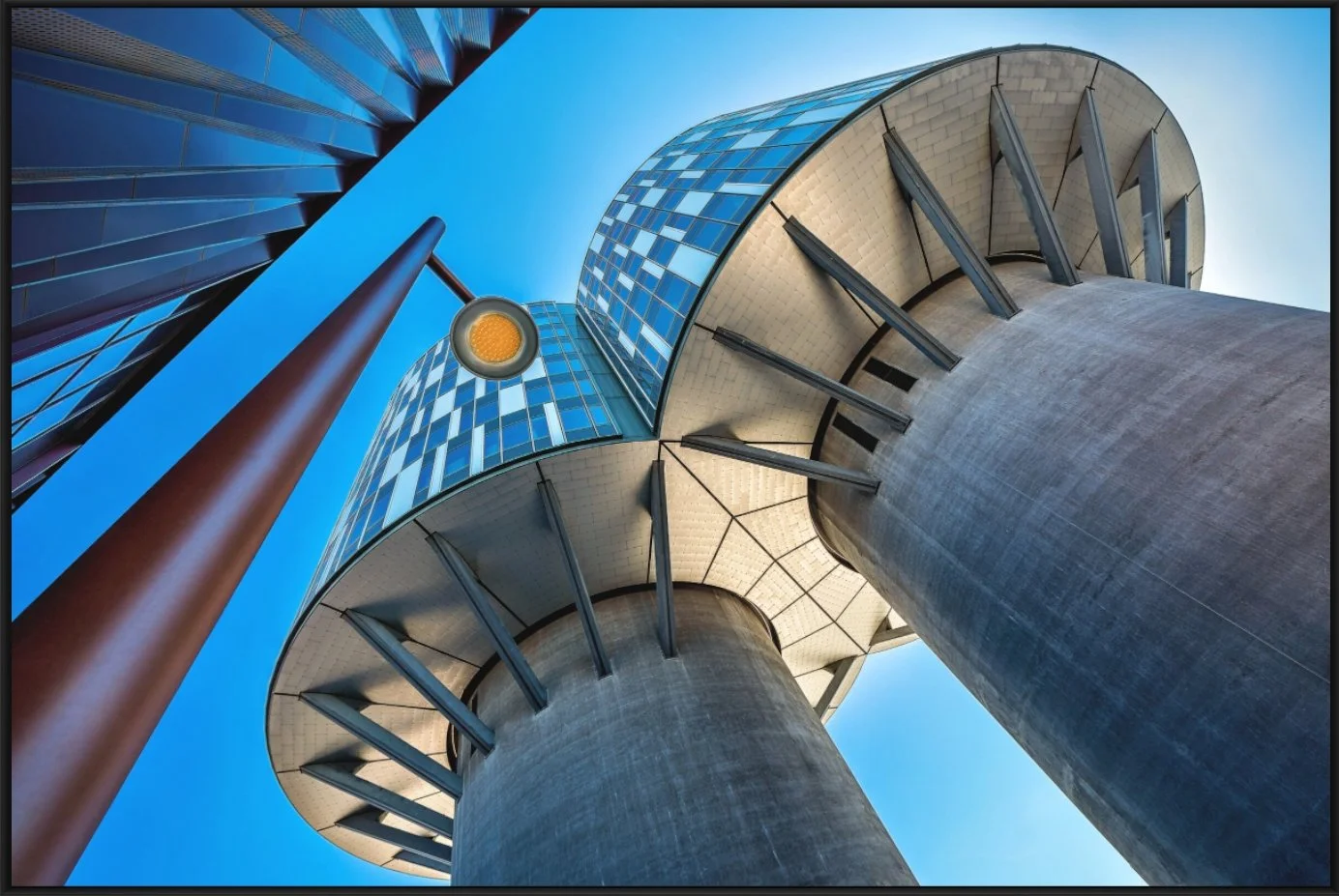  Blick von unten auf eine moderne, hohe Gebäudestruktur mit großen runden Betonpfeilern, einer geneigten Brückenkonstruktion mit Glasfassade und einer Straßenlaterne mit Gelblicht gegen den blauen Himmel.