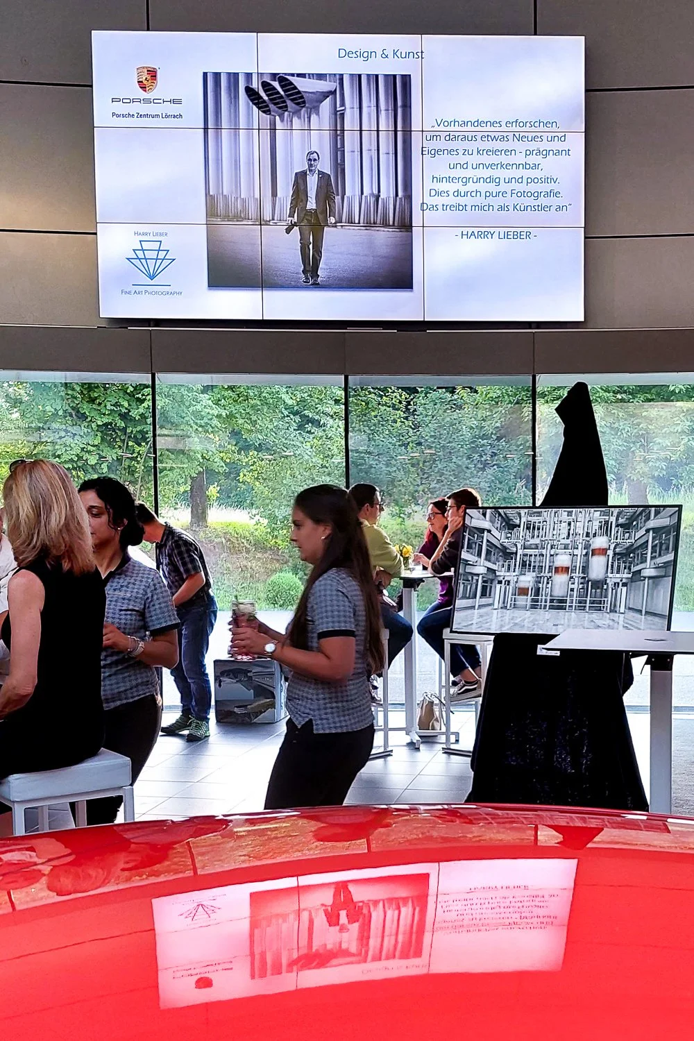 Im Veranstaltungsraum sind mehrere Menschen, die sich unterhalten und das Catering genießen. Auf einem Bildschirm ist ein Zitat eines Fotografen und ein einzelner Mann in Anzug, im Hintergrund einen Vorhang. Im Raum stehen auch ein schwarzer Aufstell