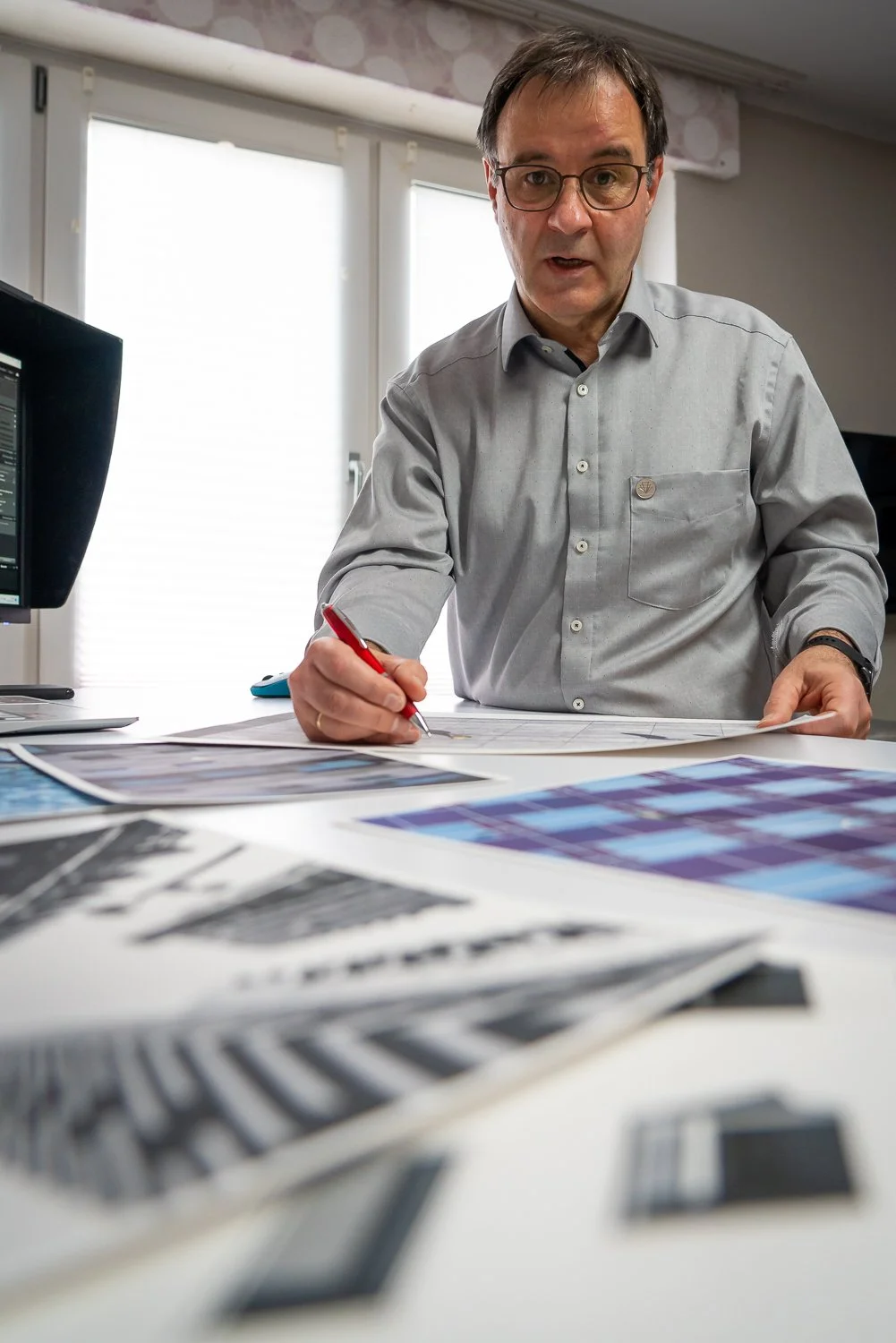 Ein Mann in einem grauen Hemd arbeitet in einem Büro am Schreibtisch, während er Skizzen und Farbfächer betrachtet.