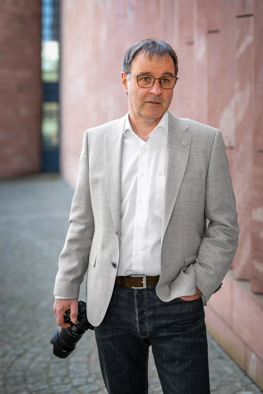 Mann in Hellblauer Anzugjacke, weißem Hemd, mit Kamera in der Hand, auf einer Straße mit rotem Backstein-Wand im Hintergrund.