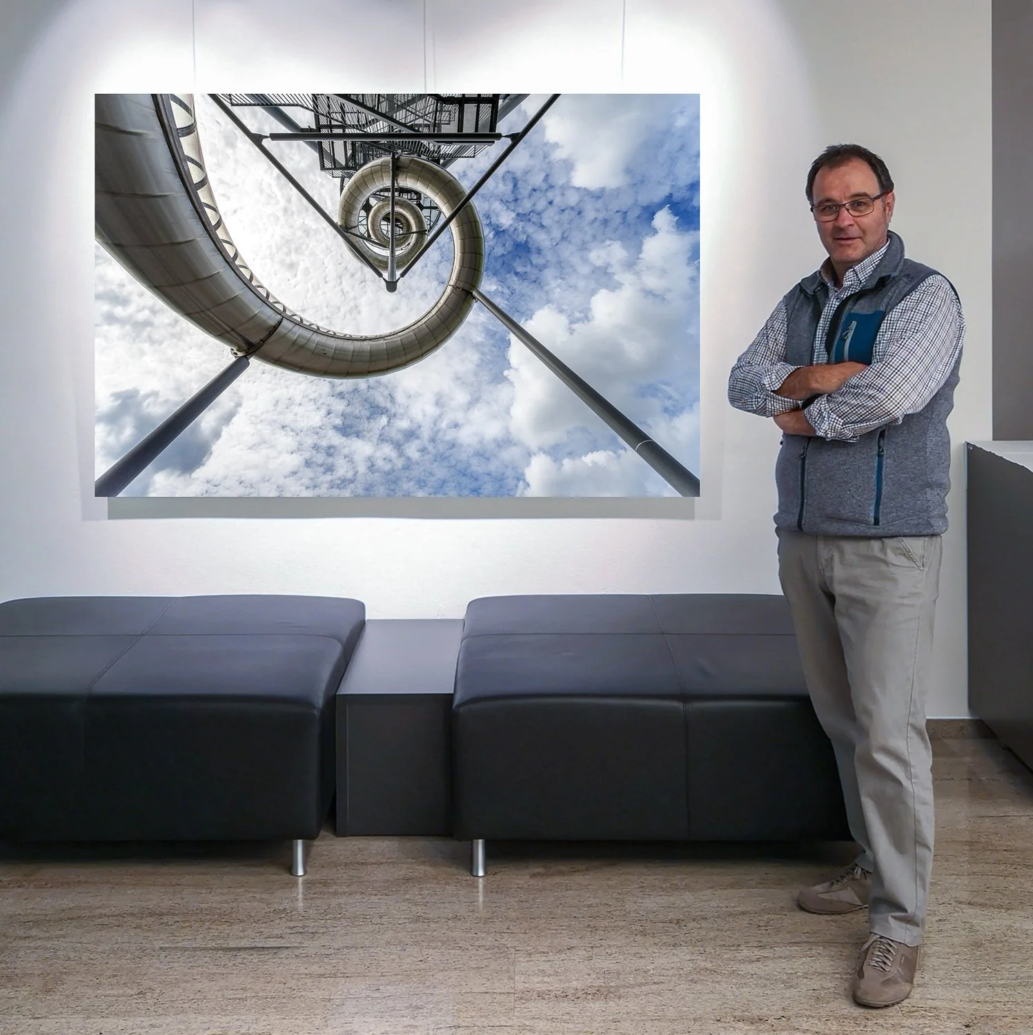Ein Mann steht in einer Galerie vor einem großen Foto, das eine spiralförmige Edelstahlrutsche, den Himmel mit Wolken und einen Aussichtsturm zeigt.
