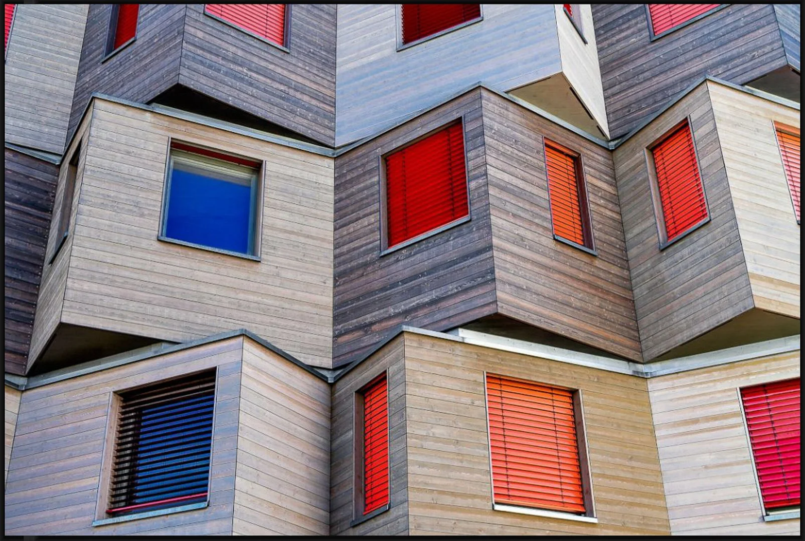 Nahaufnahme eines modernen Gebäudes mit einer Fassade aus Holz, mehreren Fenstern mit roten Jalousien und geometrischen Formen.