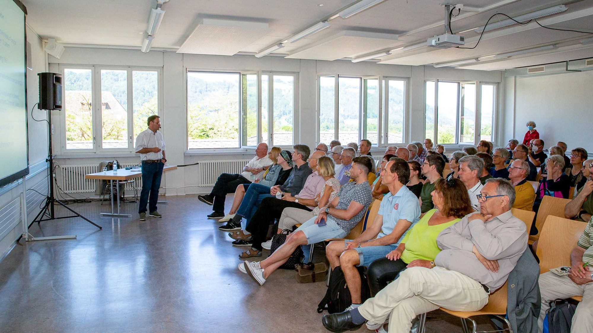 Ein Mann hält eine Präsentation vor einer Gruppe von Menschen in einem gut beleuchteten Raum mit großen Fenstern im Hintergrund.