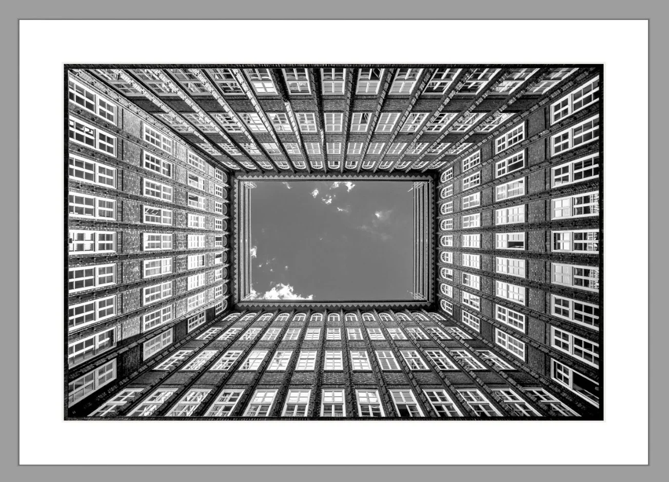 Blick nach oben in einen rechteckigen Innenhof eines mehrstöckigen Gebäudes mit vielen Fenstern, das den Himmel sichtbar macht.