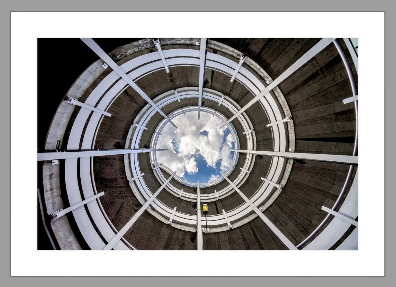 Blick nach oben in einen spiralförmigen Parkhaus-Treppenaufgang mit Sicht auf den Himmel und Wolken.