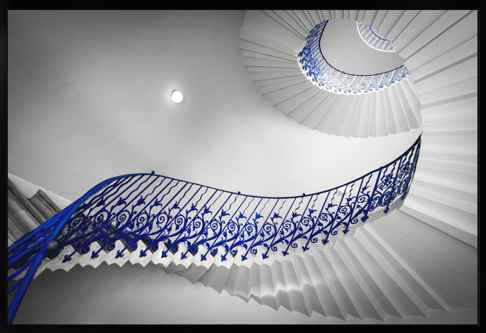 Heller weißer Treppenaufstieg mit blauer verzierter Geländer, spiralförmige Treppe nach oben, Blick von unten nach oben, mit kleiner Deckenlampe und kreisförmiger Öffnung im Deckenbereich.