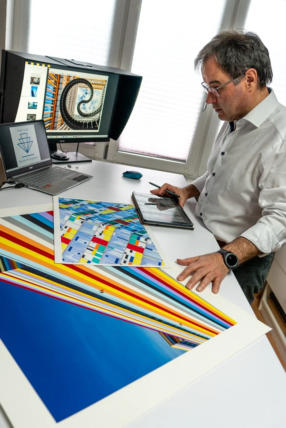 Mann arbeitet am Design von Farb- und Druckmustern mit digitalem Tablet, Computerbildschirm zeigt Spiralform, Drucke mit farbigen Geometrien liegen auf Schreibtisch