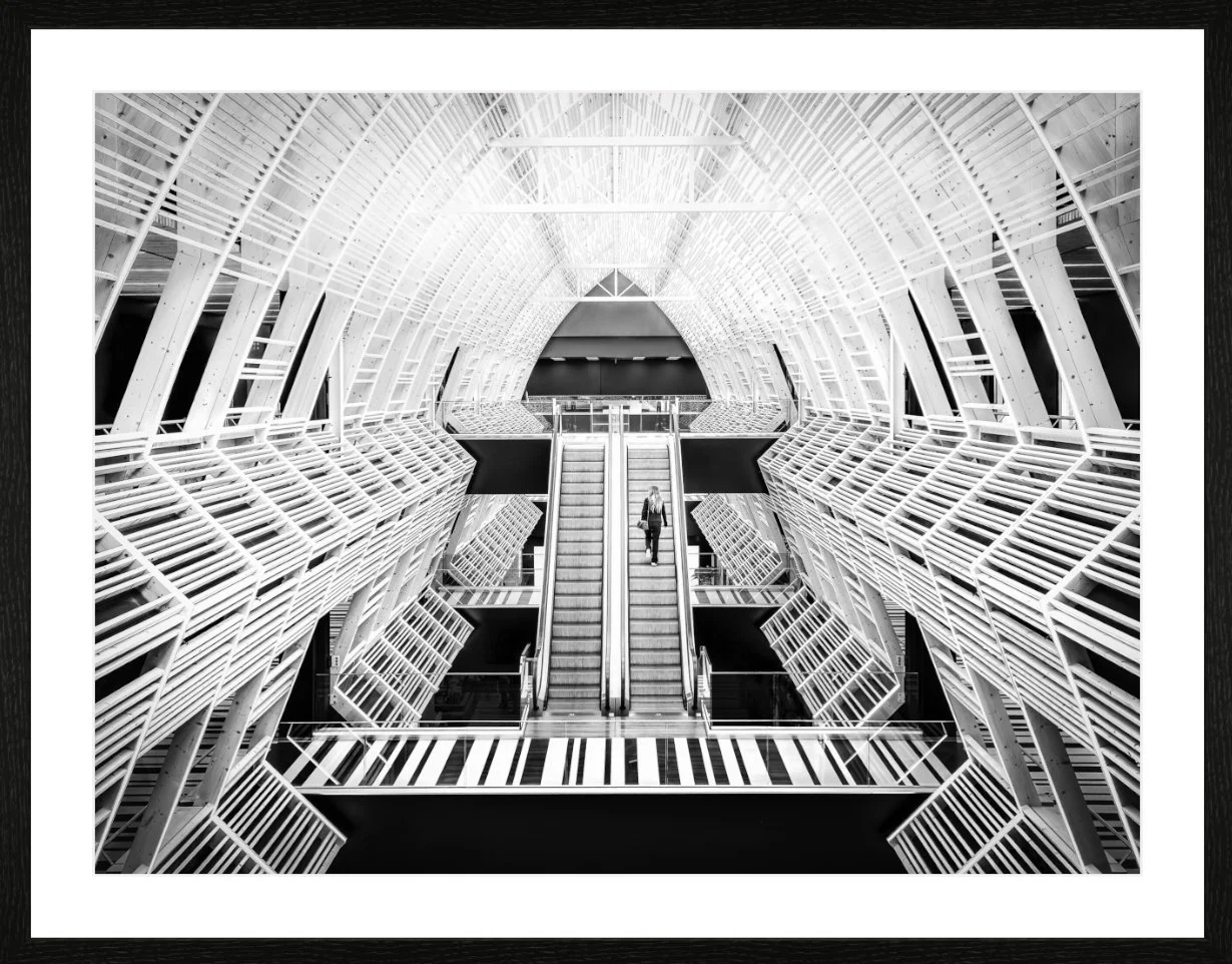 Innenansicht eines modernen, weißen Stahl-Architektur-Gebäudes mit mehreren Ebenen, Treppen und Balkonen, in dem eine einzelne Person auf einer Rolltreppe nach oben fährt.