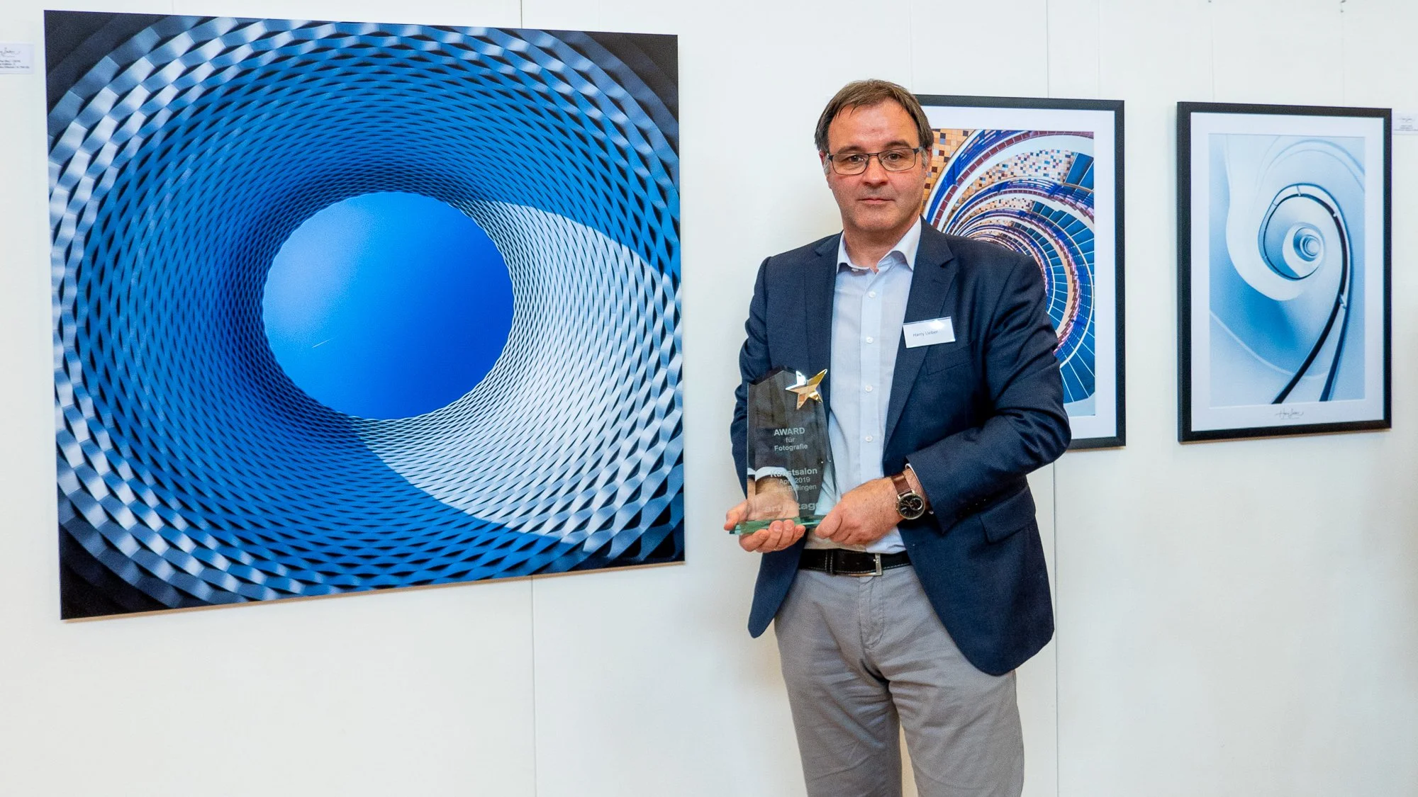 Mann in Anzügen hält einen Preis bei einer Kunstveranstaltung, umgeben von abstrakten, geometrischen Kunstwerken in blauen Tönen.