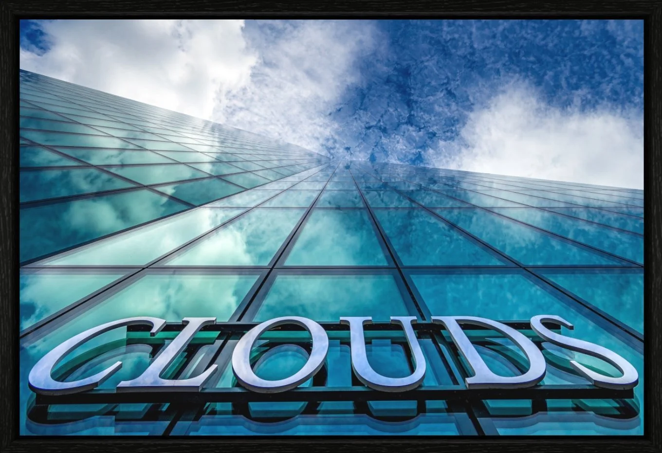 Blick nach oben auf ein modernes Glasgebäude mit der Spiegelung des Himmels und der Wolken, und einem großen Neon-Schild, das den Namen 'CLOUDS' trägt.