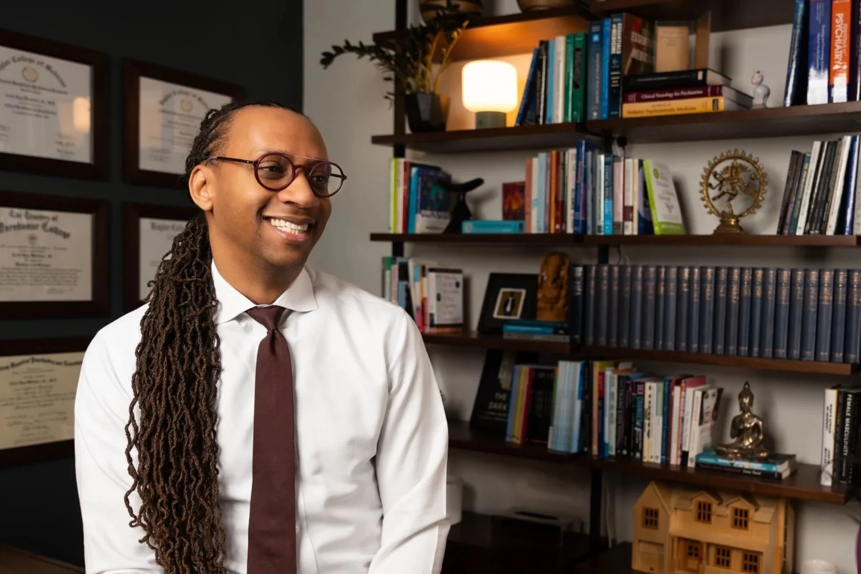 Dr. Webster, founder and CEO of Webster Clinic, smiling with glasses wearing a white shirt and maroon tie, sitting in an office with bookshelves filled with books, decor, and framed diplomas on the wall in the background.