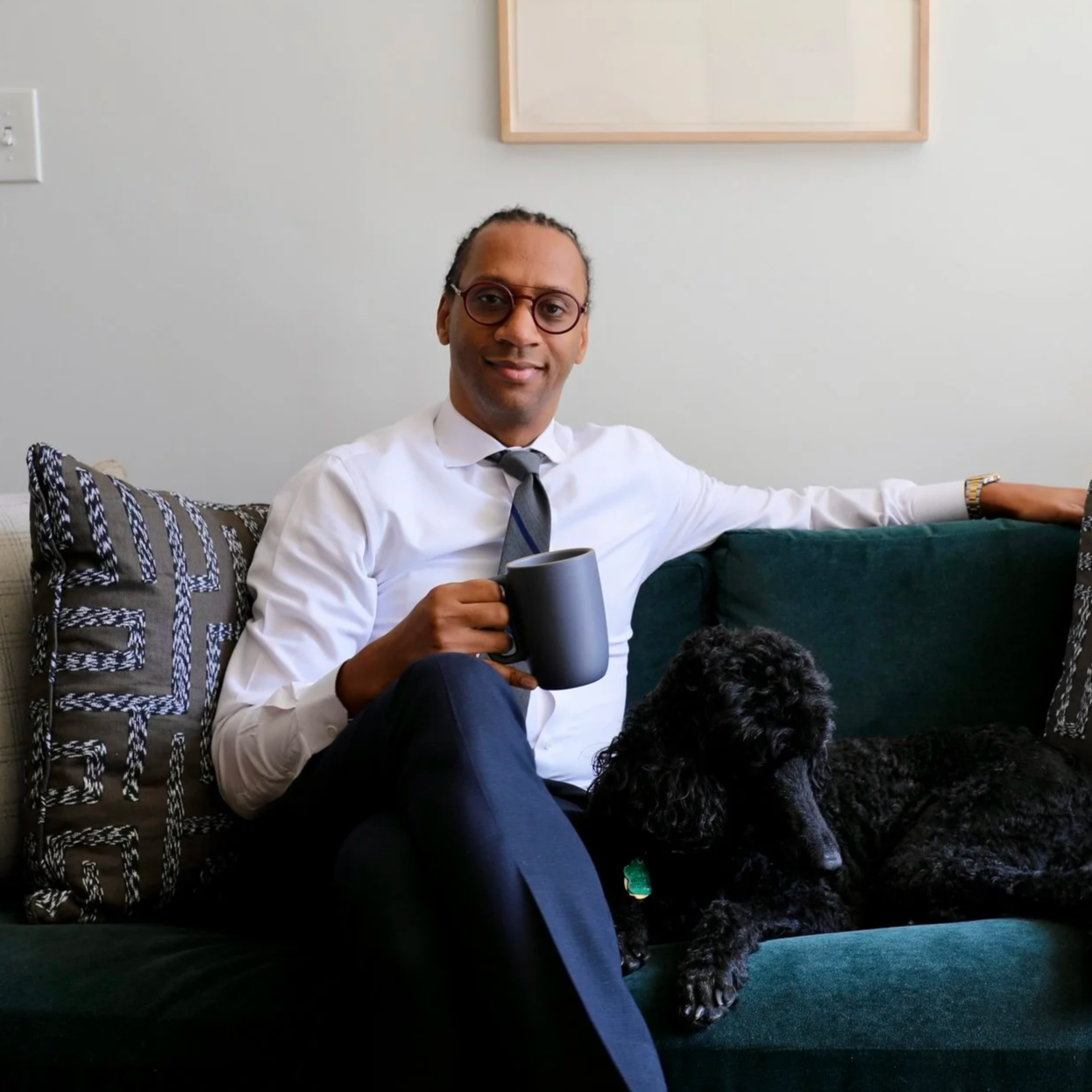 A psychiatrist in formal attire sitting on a dark green sofa with a black dog and a coffee mug, in a living room with a white wall, framed abstract art, a wooden tribal mask, a potted plant, and multiple decorative pillows.