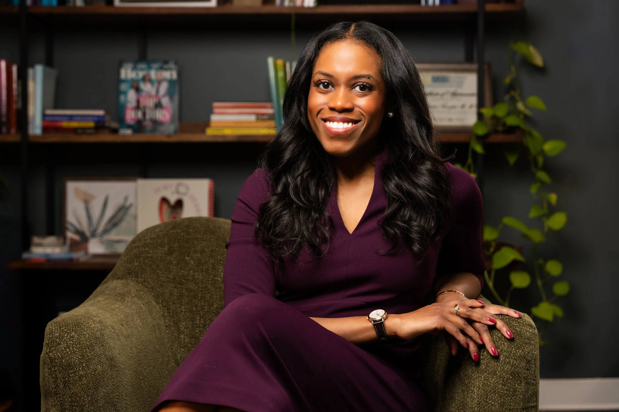 A woman with long dark hair, wearing a purple dress, smiling and sitting on an olive green armchair in a room with a bookshelf and a potted plant in the background.
