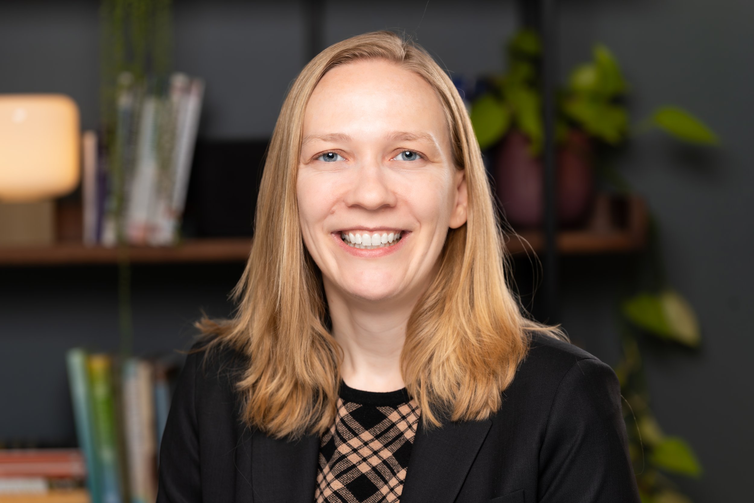 Portrait of a smiling woman with shoulder-length blonde hair, wearing a black blazer and a patterned top, sitting in an office or library setting with shelves of books and plants in the background. Child and adolescent and adult psychiatrist.