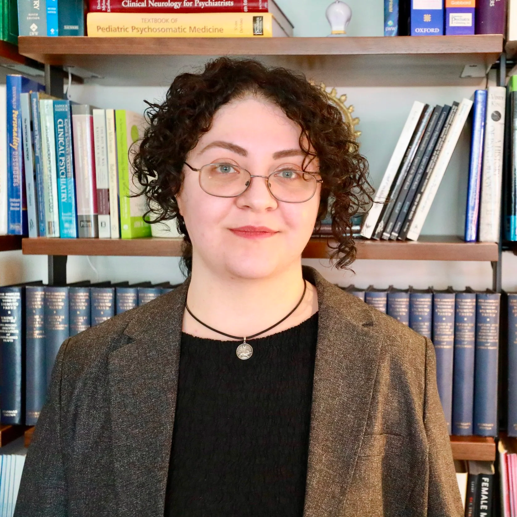 A woman with short curly black hair and glasses, wearing a black top, a white button-up shirt, and a black choker with a red pendant, standing against a plain light-colored wall.