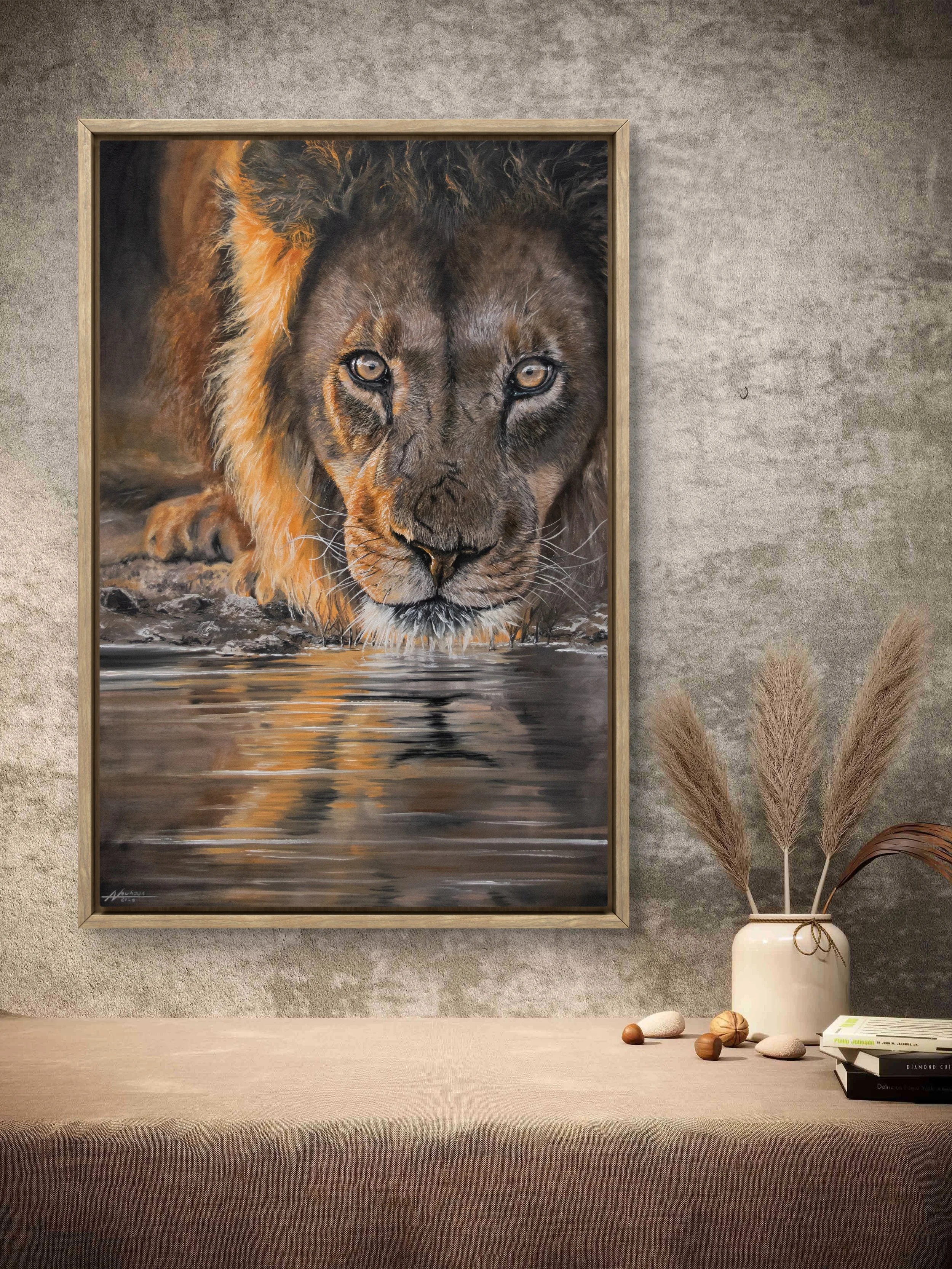 Painting of a lion drinking water, hung on a wall in a room with neutral decor, next to a vase with dried grasses and books on a table.
