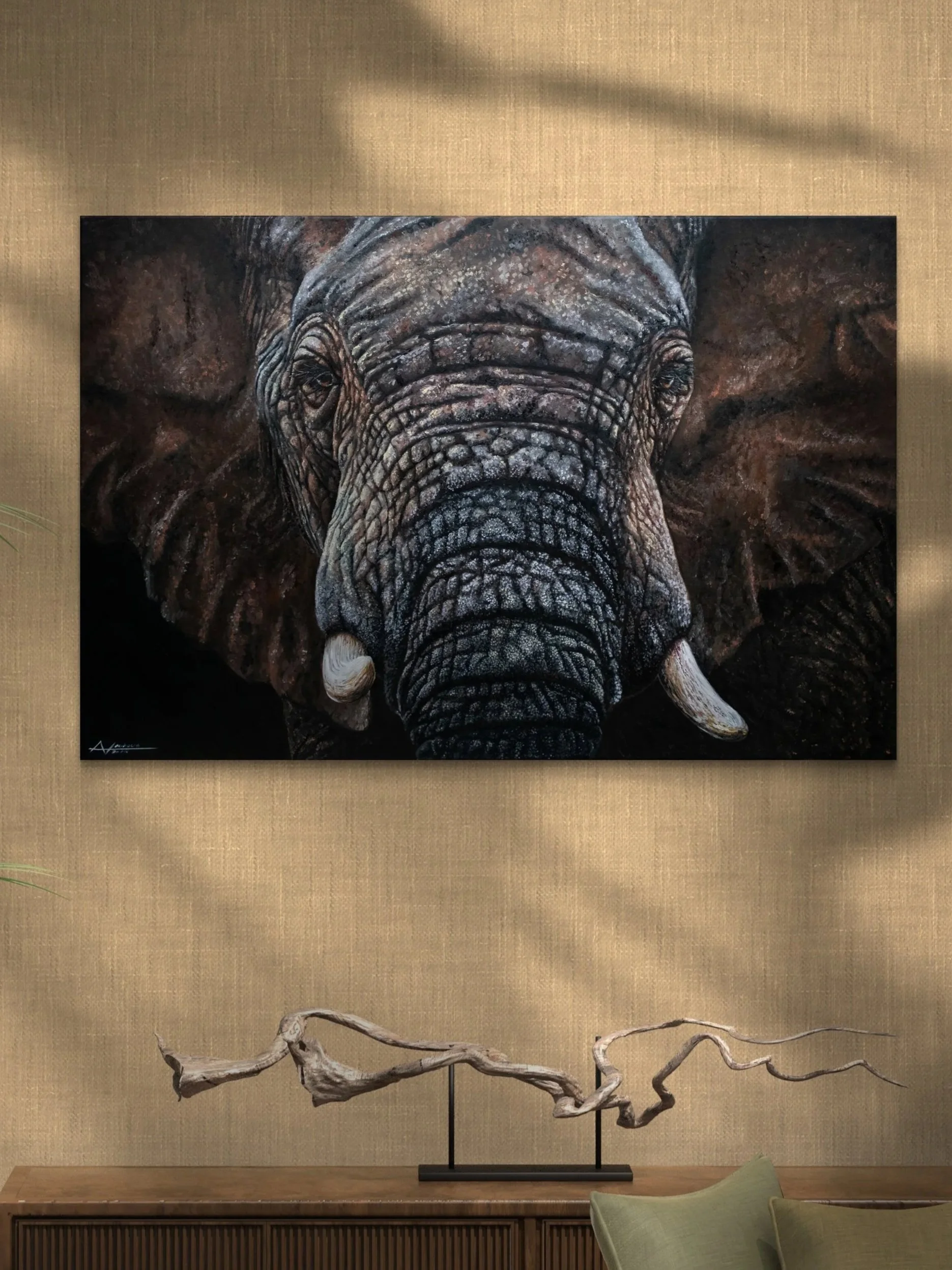 A painting of an elephant's face hung on a beige wall above a wooden console with a twisted, driftwood sculpture and a green cushion on a chair in the foreground.