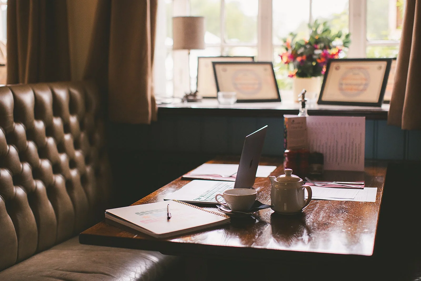 A laptop sits open next to a notebook and a pot of tea on a wooden dining table in front of a 16th century bay window
