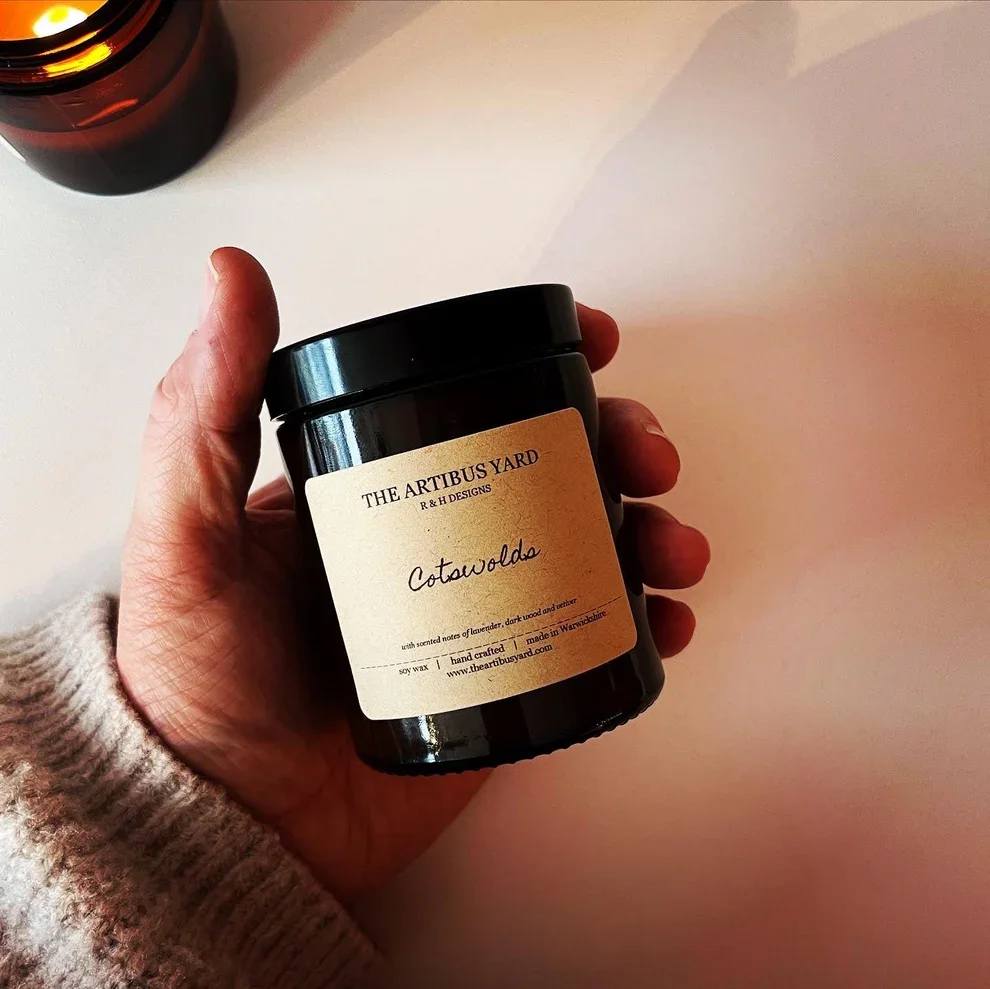 A hand holds an artisan unopened candle jar called Cotswolds made by The Artibus Yard with scents of lavender and dark wood.