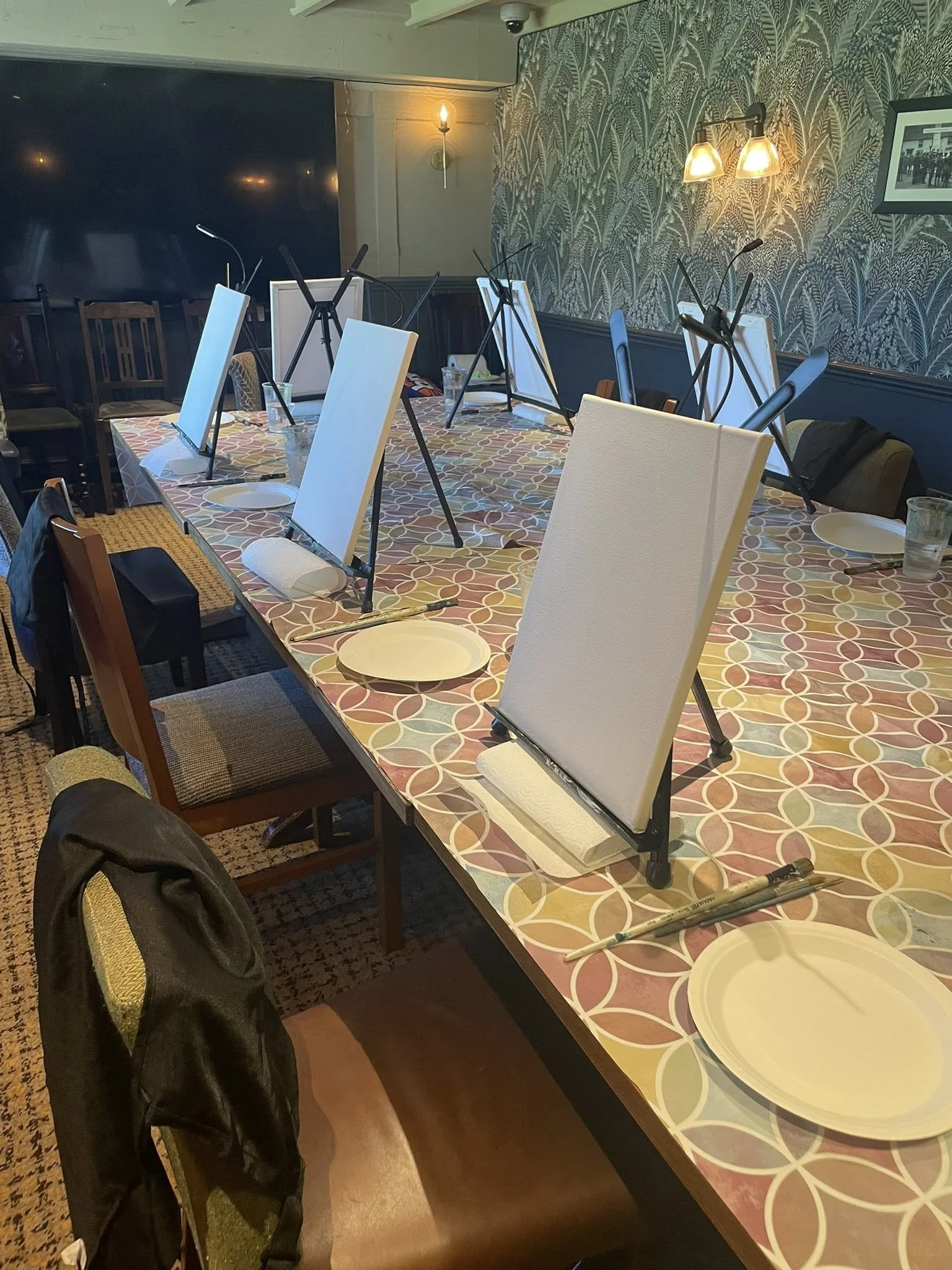 6 small canvases sit on tripods on a communal table with paint palettes and brushes waiting to be used.