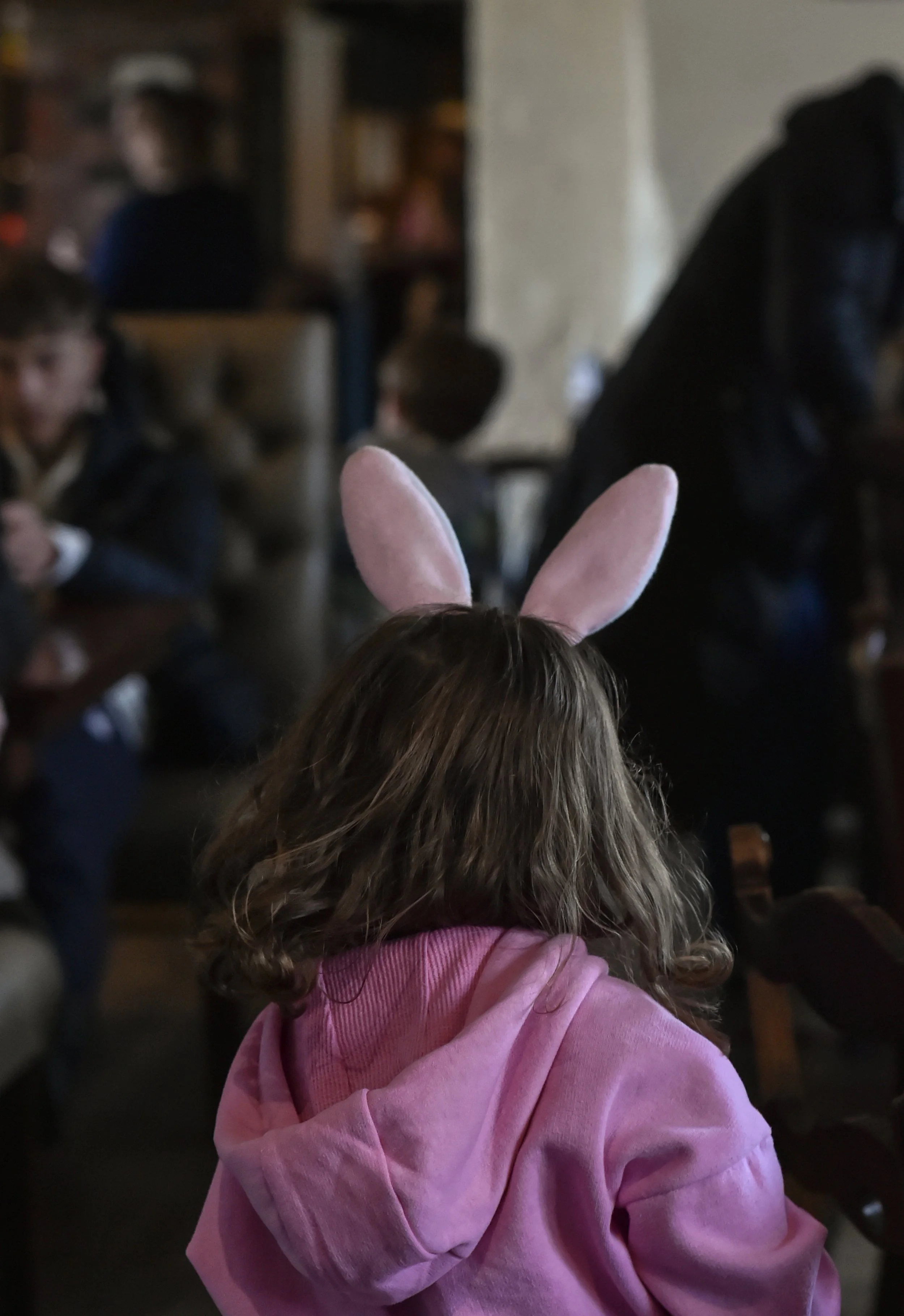 A little girl from behind running into a busy room wearing a pink hoodie and bunny ears.