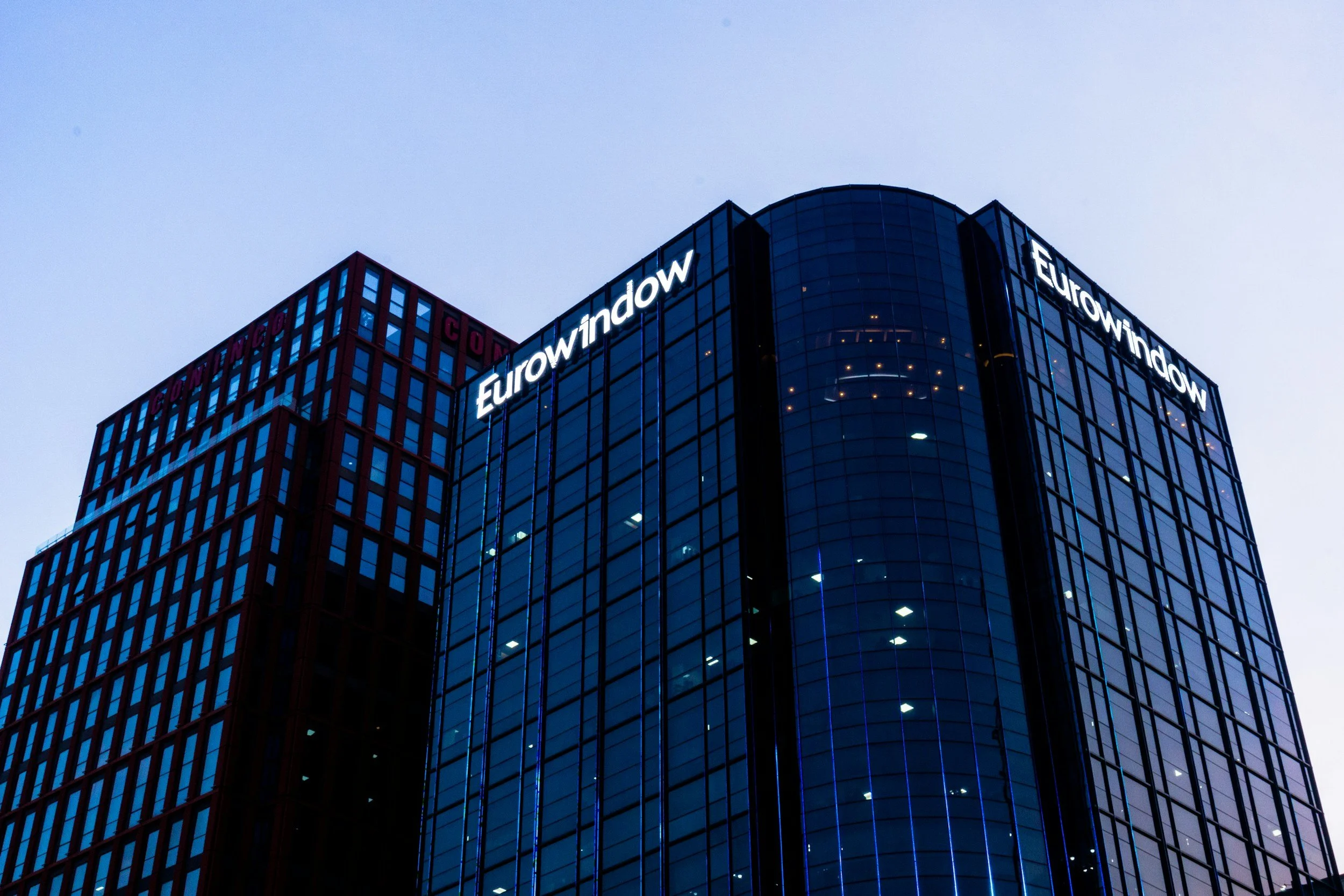 Nighttime view of a modern glass office building with illuminated Eurowindow signs at the top.