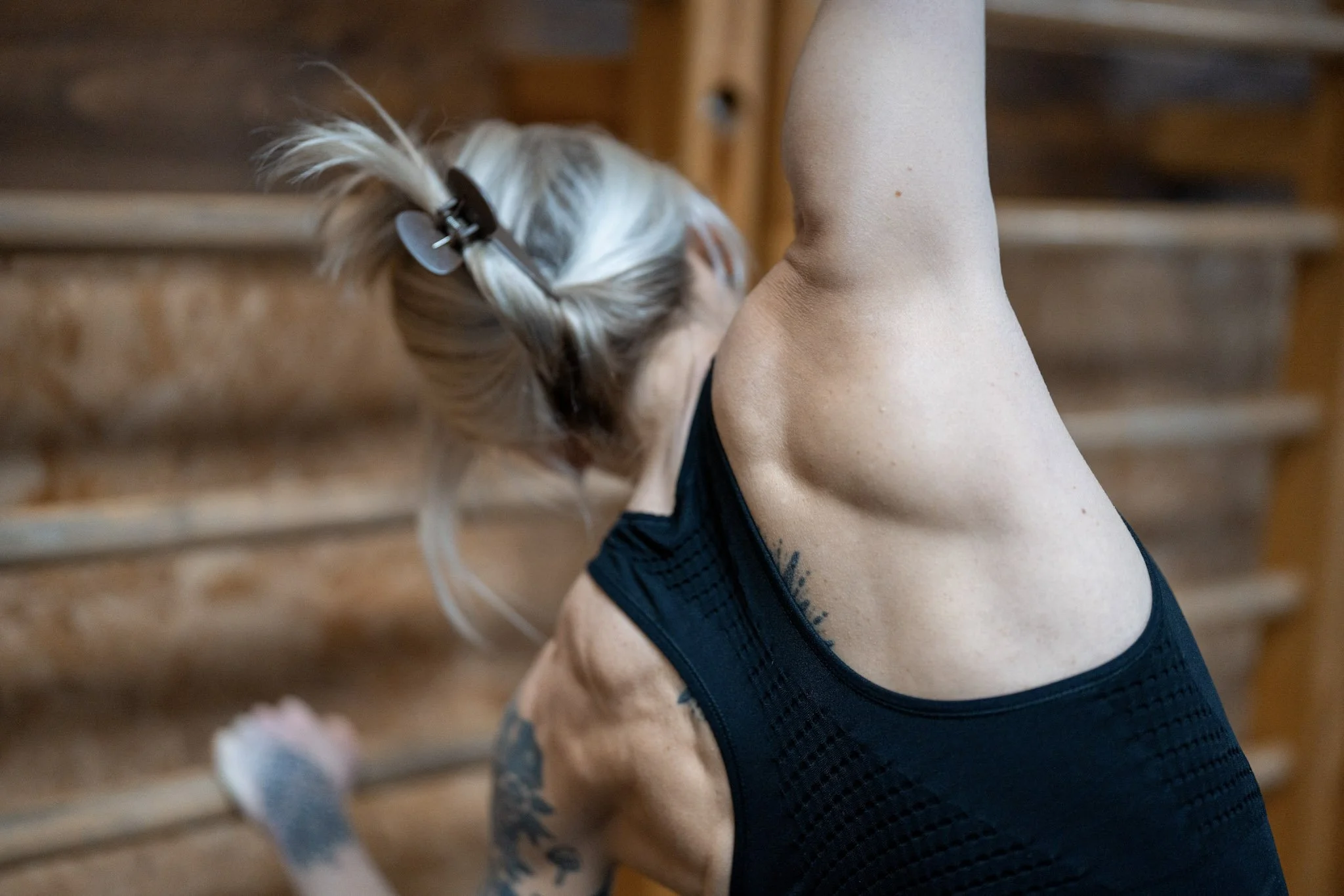 A woman with blonde hair tied with a hair clip, wearing a black athletic top, stretching her right arm overhead in a gym or fitness space with wooden equipment in the background.
