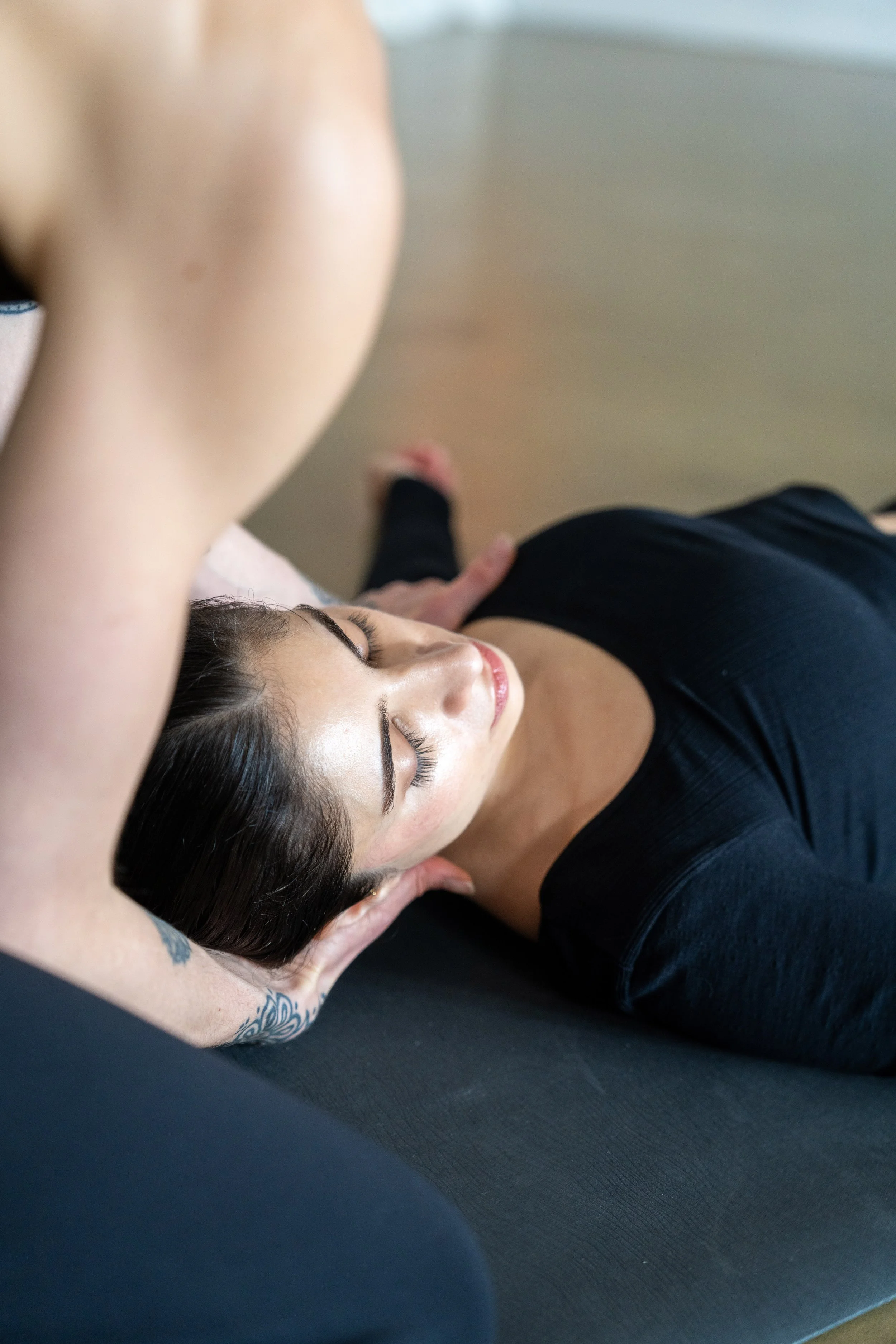 A woman is lying on her back on a massage table with her eyes closed, receiving a head massage from a therapist whose hands are on her head.