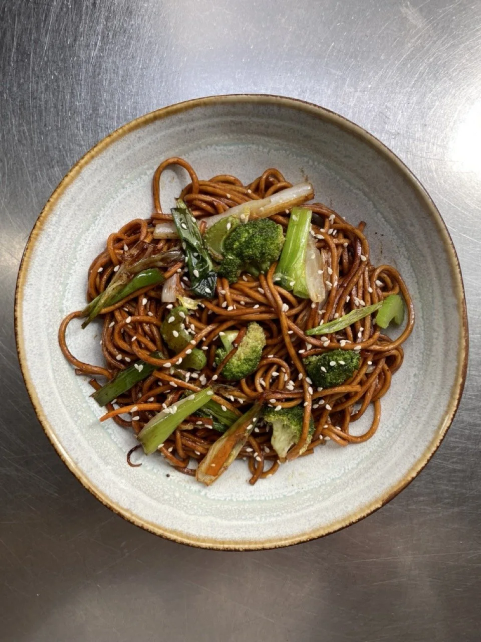 Plat de nouilles sautées avec légumes, notamment brocoli, oignons verts et carottes, garni de graines de soja ou de sésame.