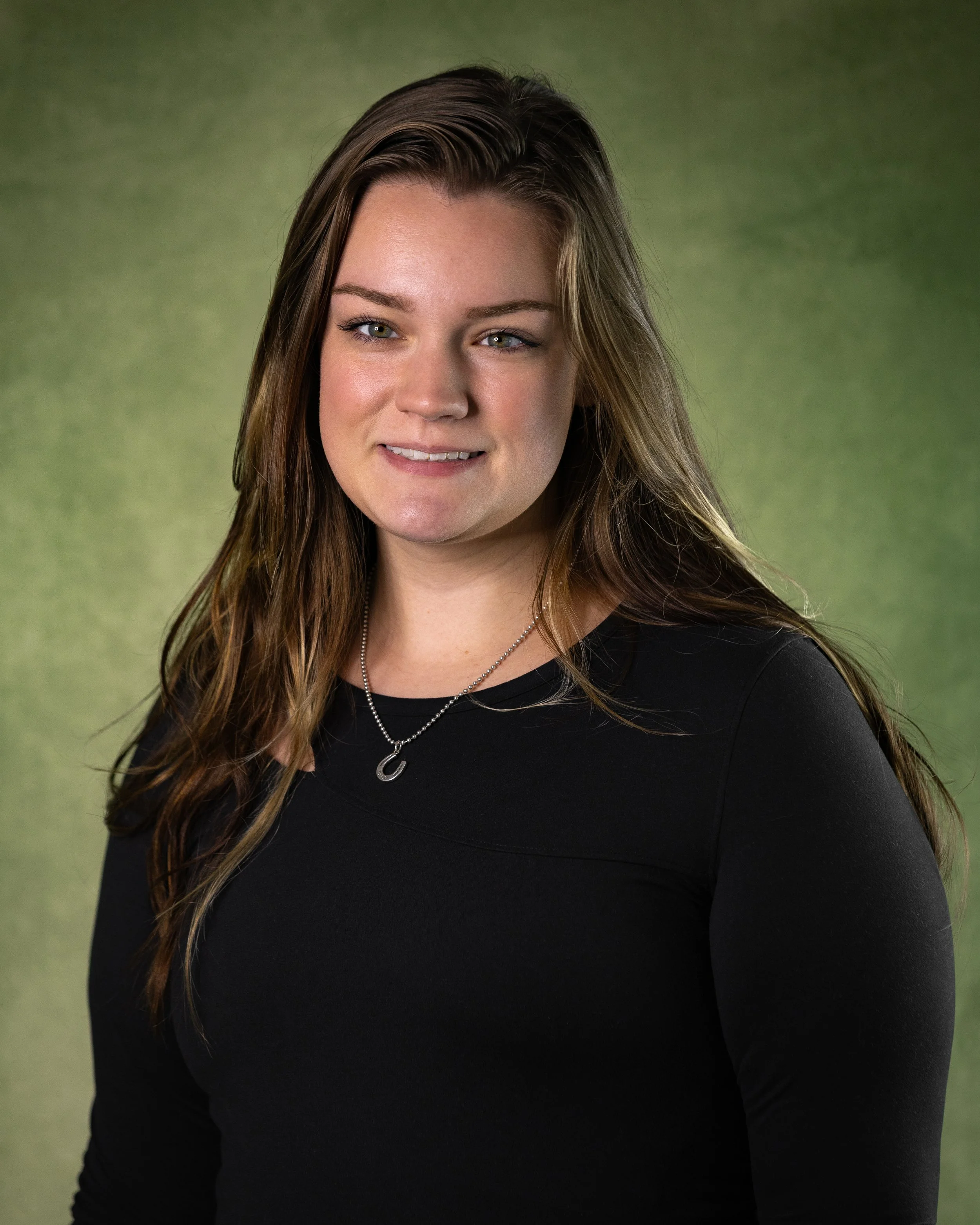A young woman with long brown hair, wearing a black top and a silver necklace with a 'C' pendant, smiling softly in front of a green background.