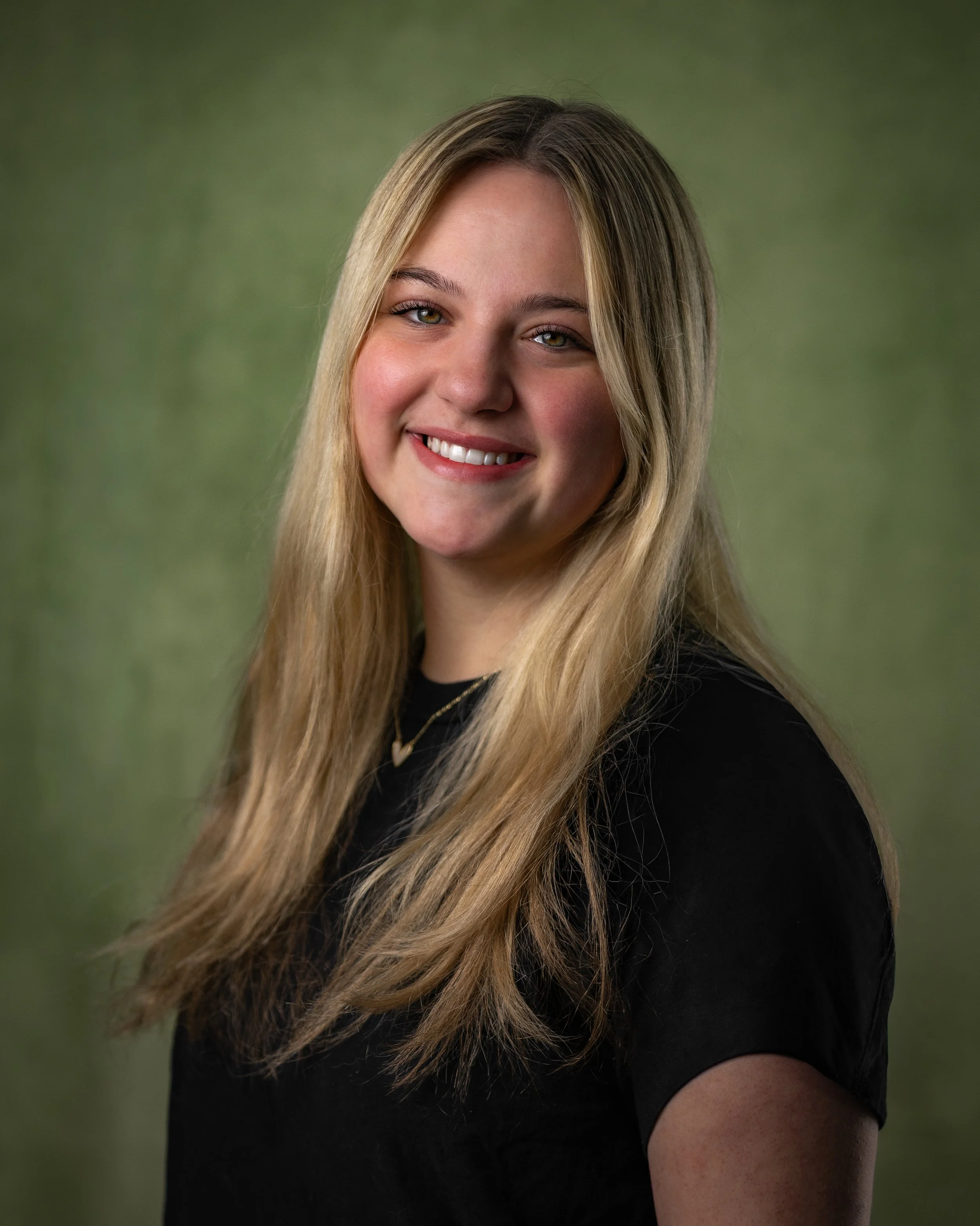 A young woman with long blonde hair smiling, wearing a black shirt, against a green background.