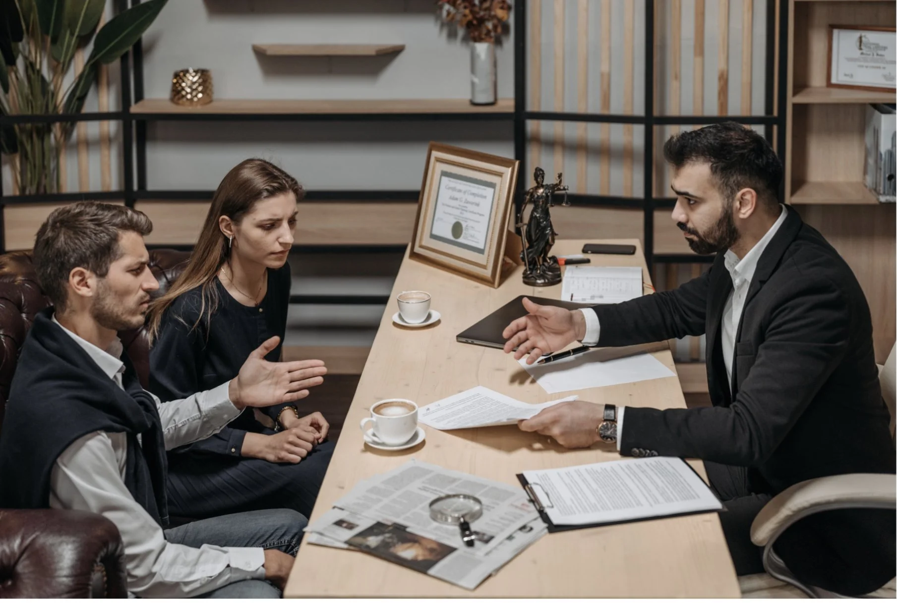 A man and a woman on the left side of the table are engaged in a serious conversation with a man on the right side, who appears to be a professional or counselor. The setting is an office with documents, coffee cups, a framed certificate, and decorative items on the desk.