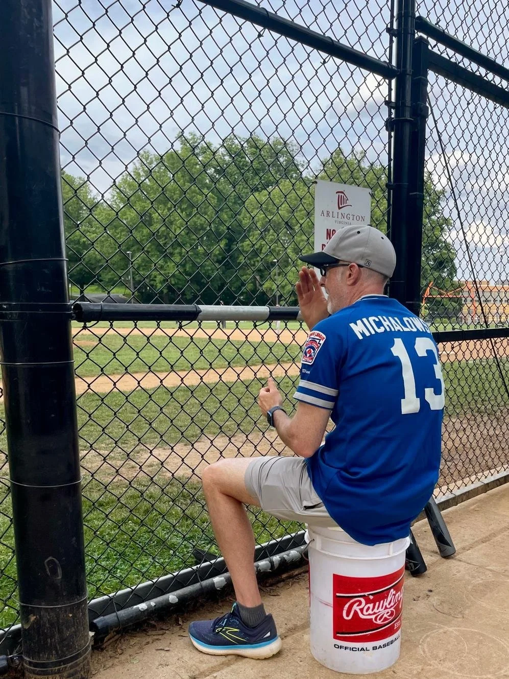 A man in a blue sports jersey its on a white baseball bucket labeled 'Rawlings' outside a baseball field.