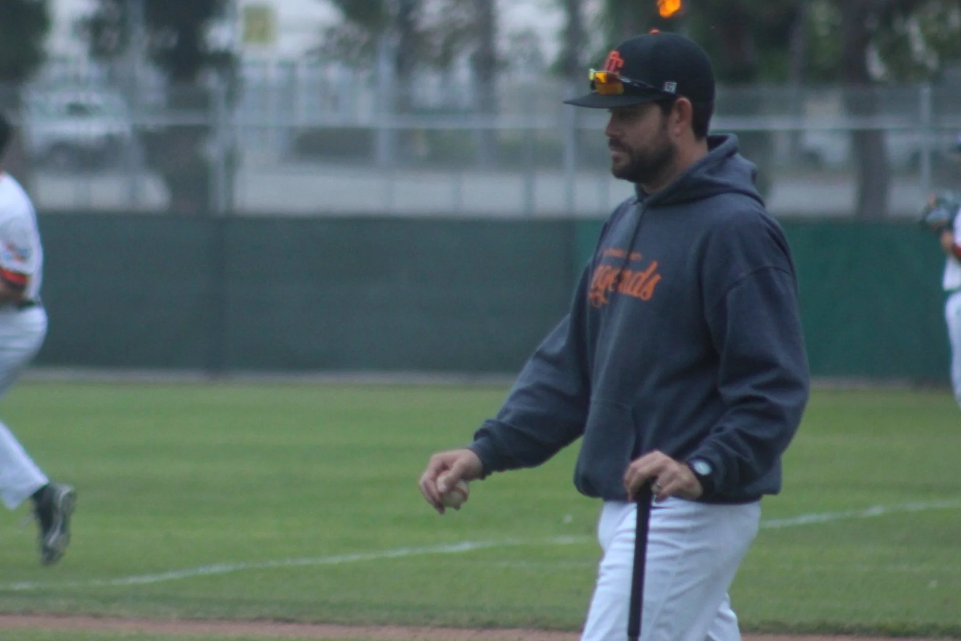 A man in a black hoodie and white baseball pants stands on a baseball field, holding a bat, with other players in the background.