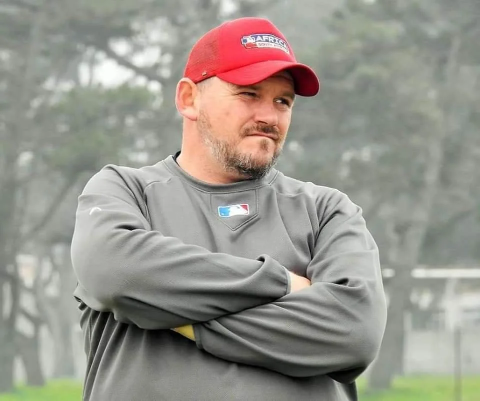A coach wearing a red baseball cap and gray sweatshirt standing at a baseball field.