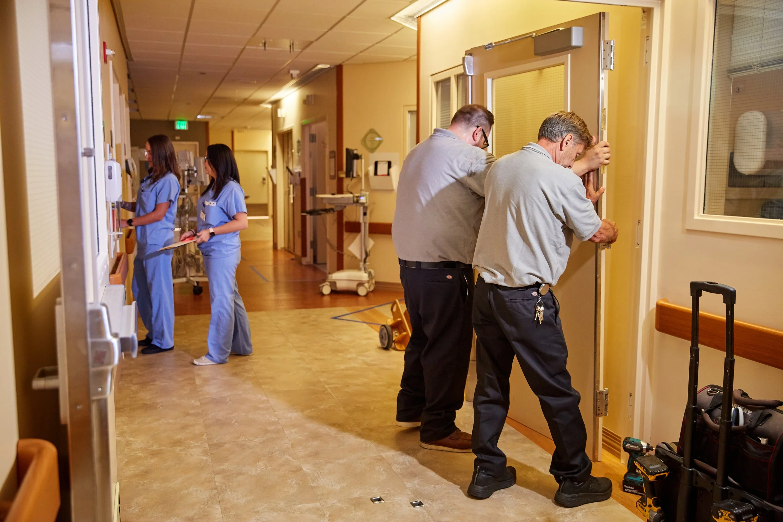 Two men installing a door in a hospital hallway, with two nurses working in the background.