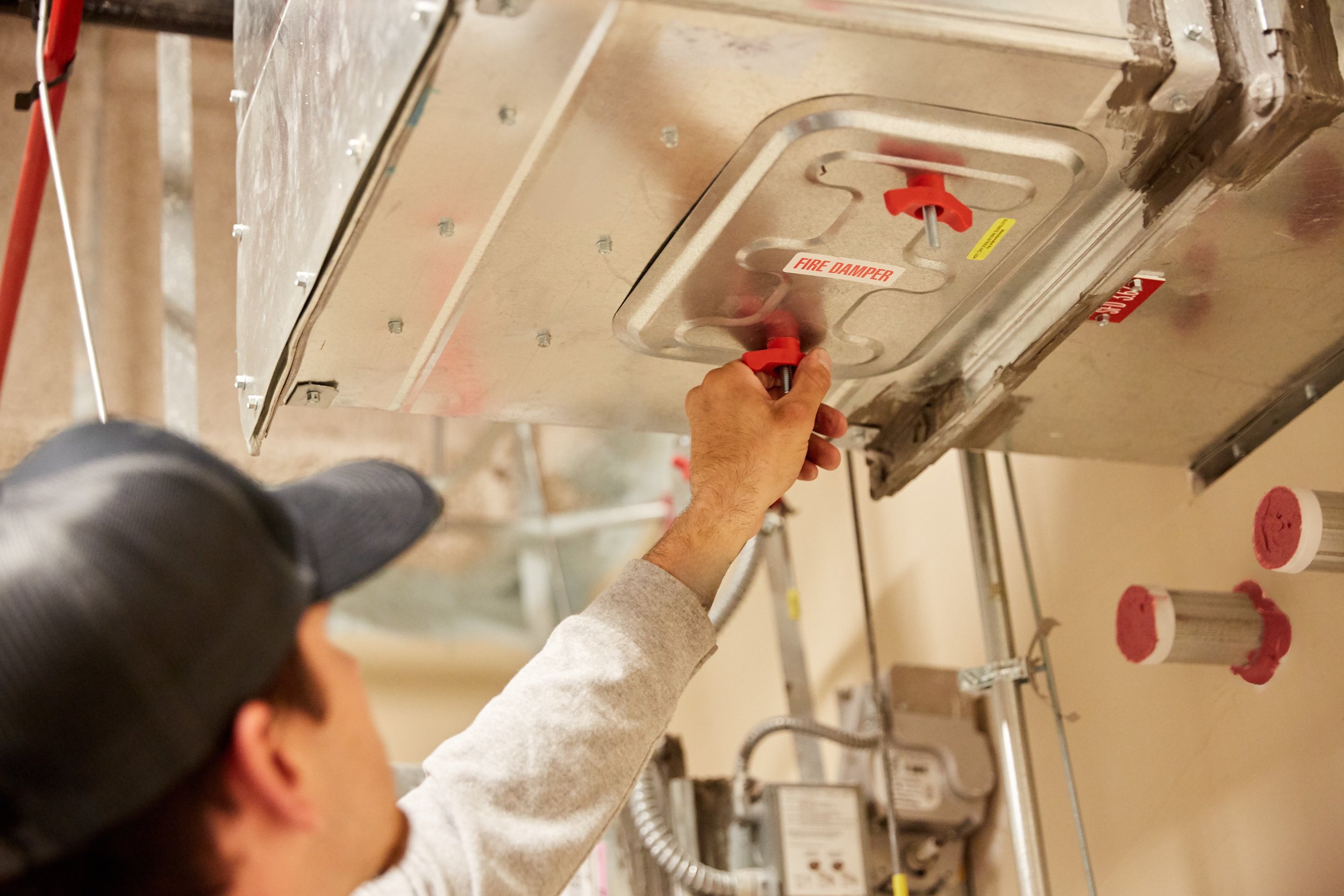 A person wearing a gray long sleeve shirt and a dark baseball cap adjusting a fire damper on an industrial HVAC system ceiling.